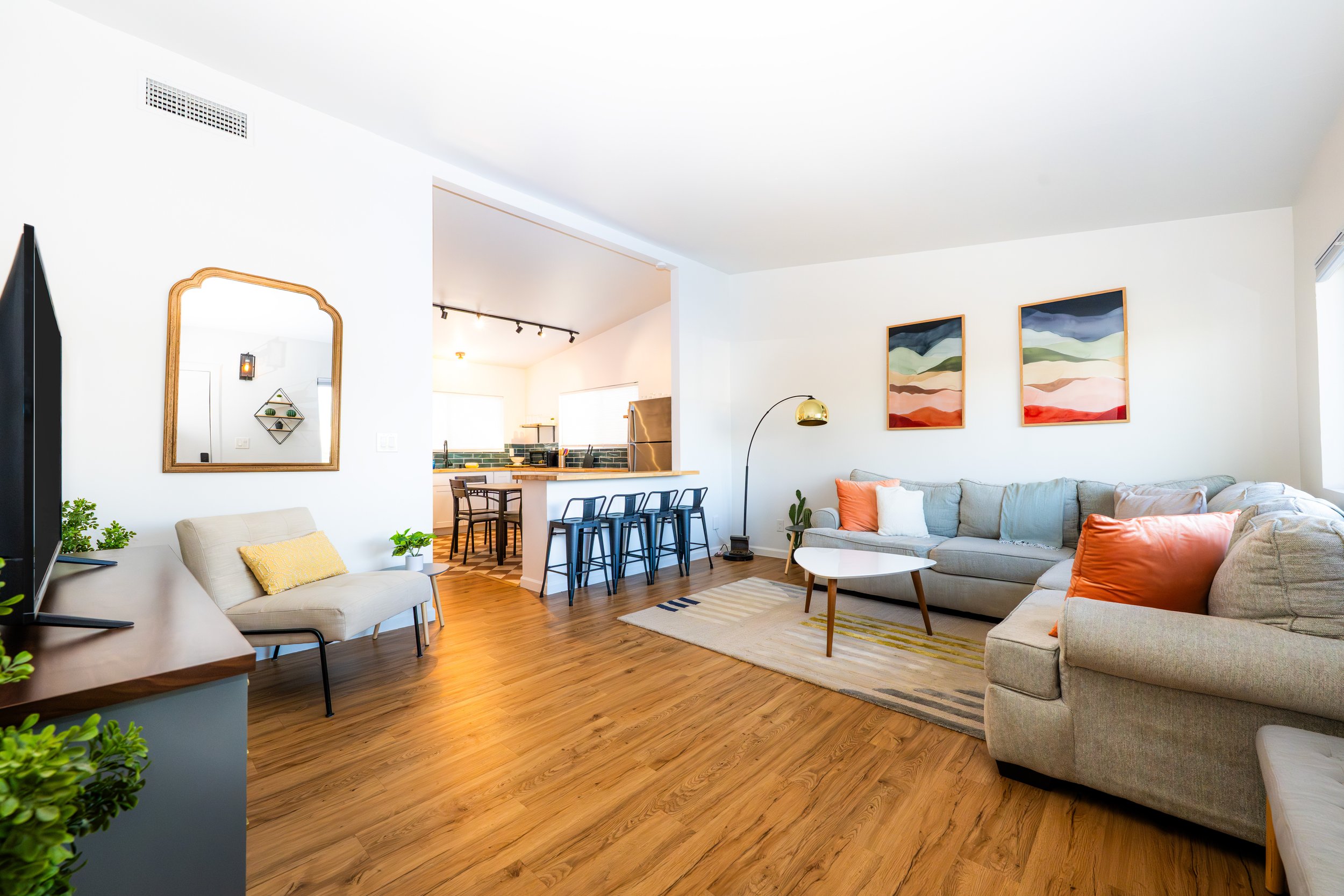 Bright living room with gray sectional sofa, pastel and orange throw pillows, abstract landscape artwork, wooden coffee table, and a gold floor lamp. Open kitchen and bar stools in the background.