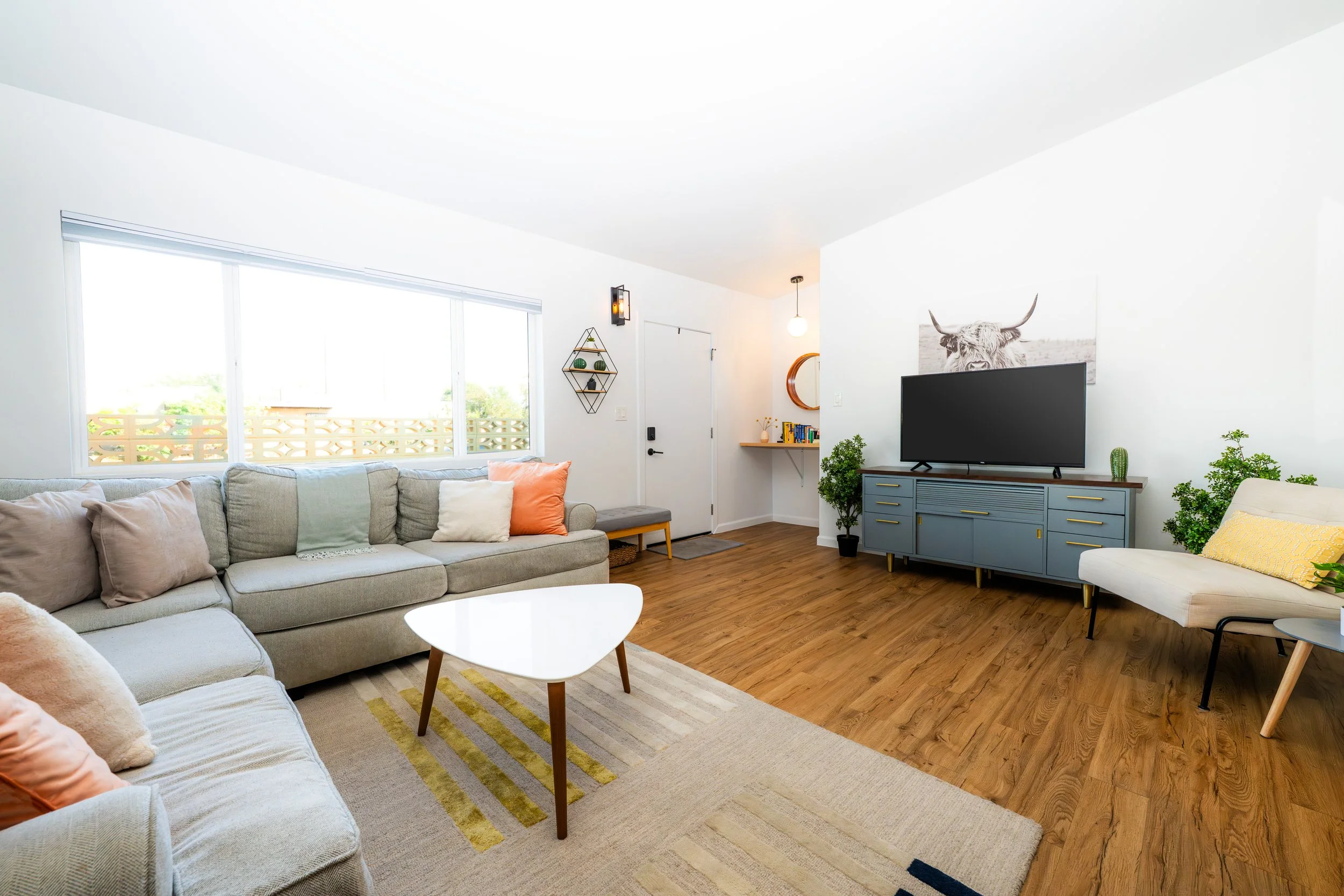 Bright living room with large window, beige sectional sofa with colorful pillows, white oval coffee table, gray and yellow striped rug, blue TV stand with a flat-screen TV, artwork of a cow, plants, and cozy decor.
