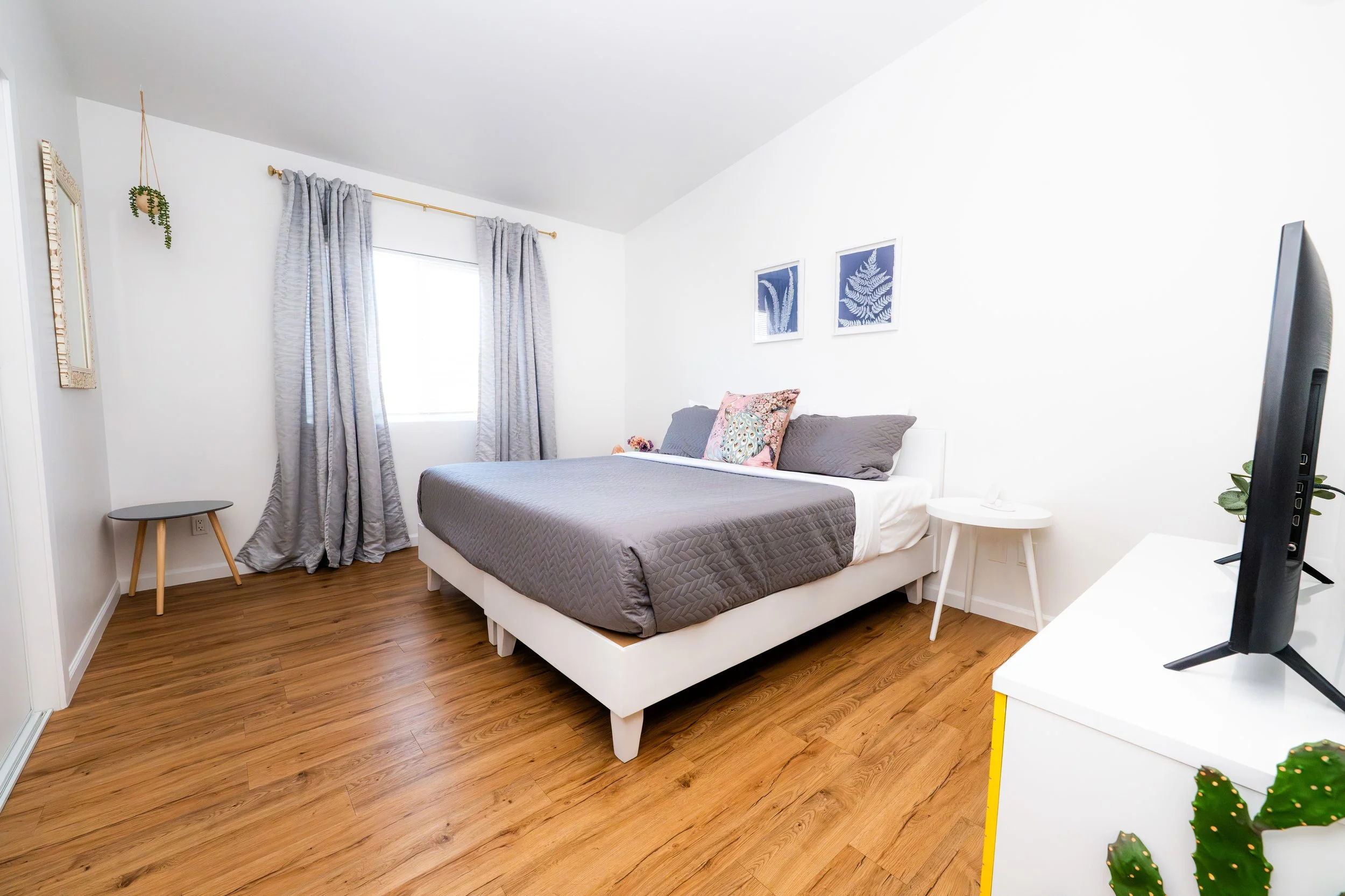 A modern bedroom with a white bed frame, gray quilt, and decorative pillows. Light wood flooring, a window with gray curtains, two framed botanical prints on the wall, a small side table, and a white dresser with a flat-screen TV.