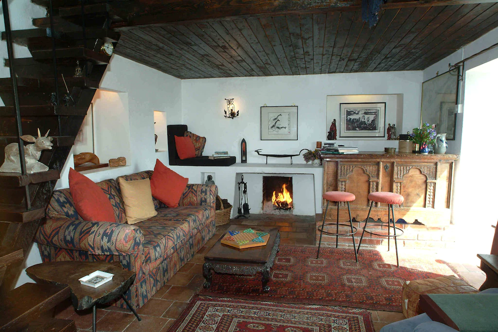 Cozy living room with a colorful patterned sofa, wooden coffee table, fireplace, vintage wooden sideboard, bar stools, and decorative art on the white walls, with a dark wood ceiling and stairs to the left.