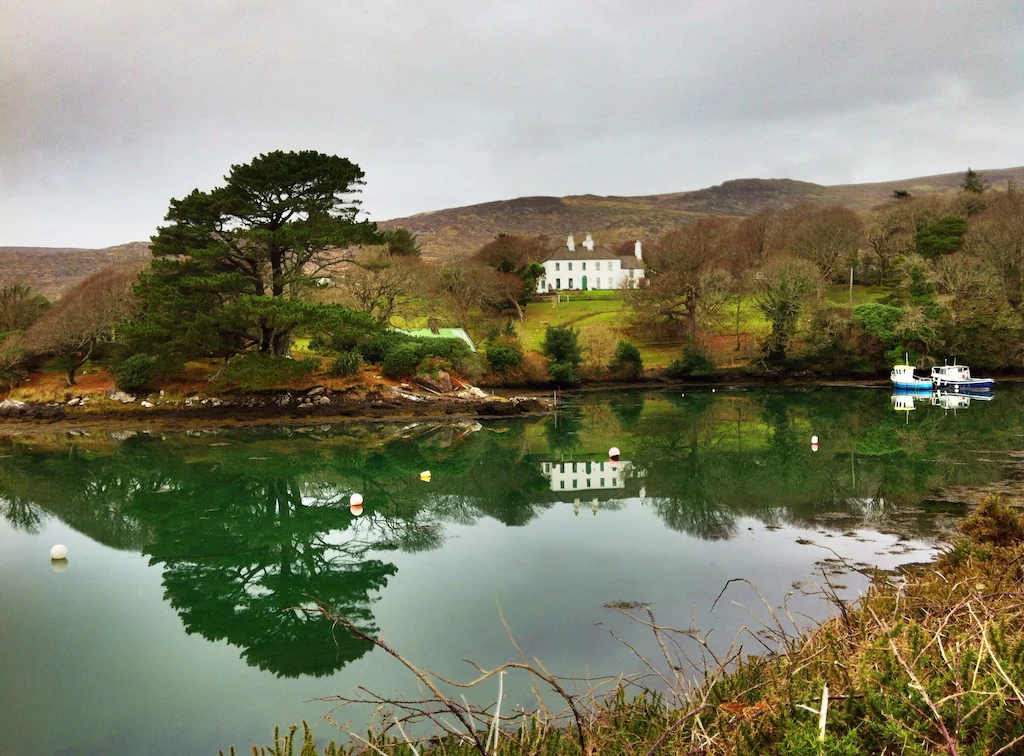 A scenic view of a body of water reflecting a white house, several trees, and boats on the shore, with hills and a cloudy sky in the background.