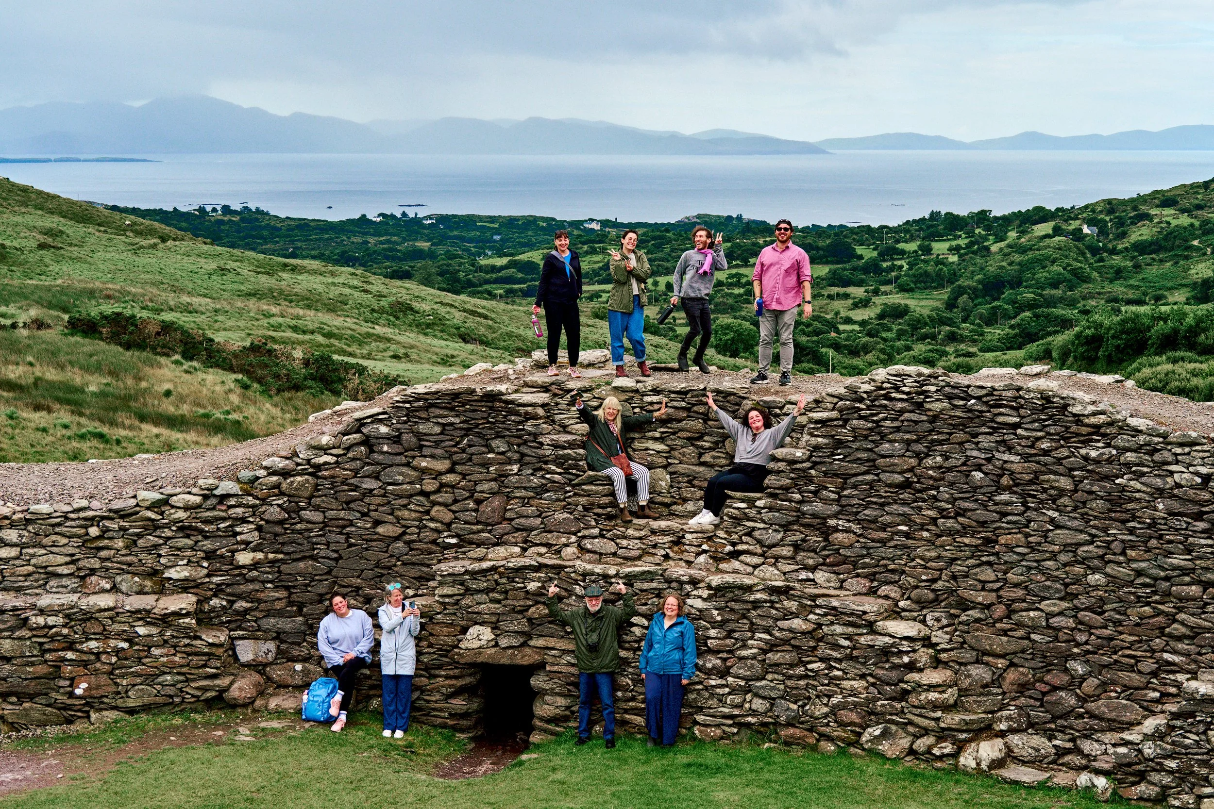 A group of people posing on and around ancient stone ruins outdoors with green hills and a body of water in the background.