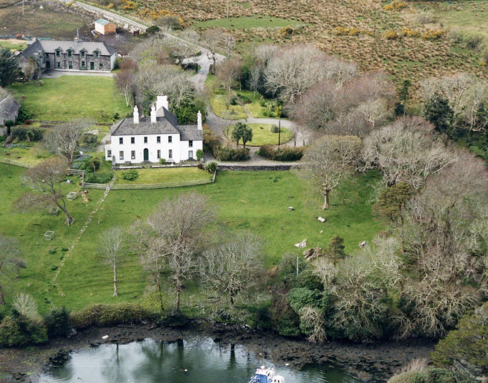 Aerial view of a large white house with a dark roof, surrounded by a manicured lawn, trees, and a pond in the foreground.
