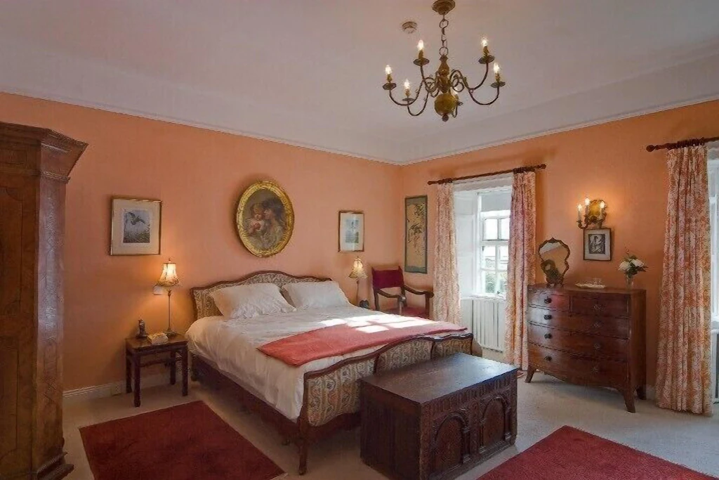 A cozy bedroom with peach walls, a large bed with white linens and a red blanket, wooden furniture including a wardrobe, chest of drawers, and bedside tables, along with a chandelier and wall-mounted lights, decorated with framed artwork and floral c