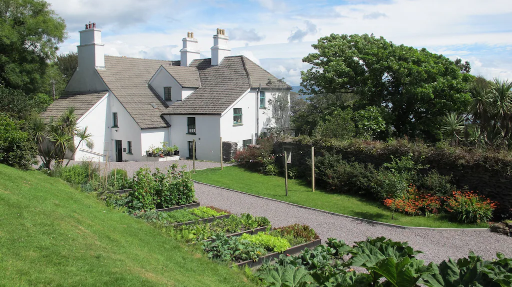 White house with multiple chimneys surrounded by lush green trees and a garden with raised vegetable beds and colorful flowers.