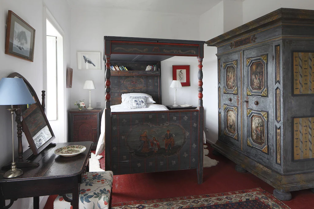 A vintage bedroom with dark wooden furniture, a four-poster bed, a large armoire, a small desk, and various framed artwork on white walls.