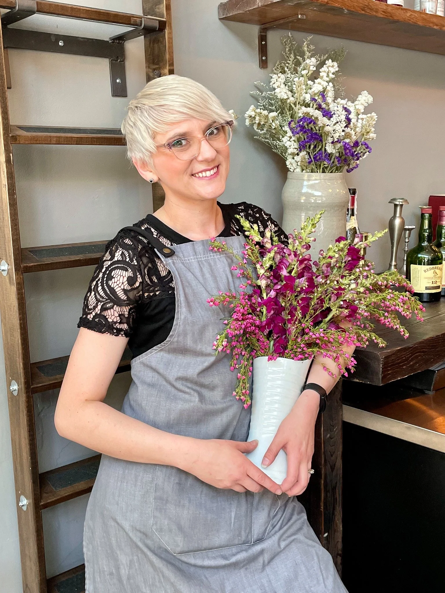 A woman with short blonde hair and glasses holding a white vase filled with pink and purple flowers, smiling in a kitchen or bar area.