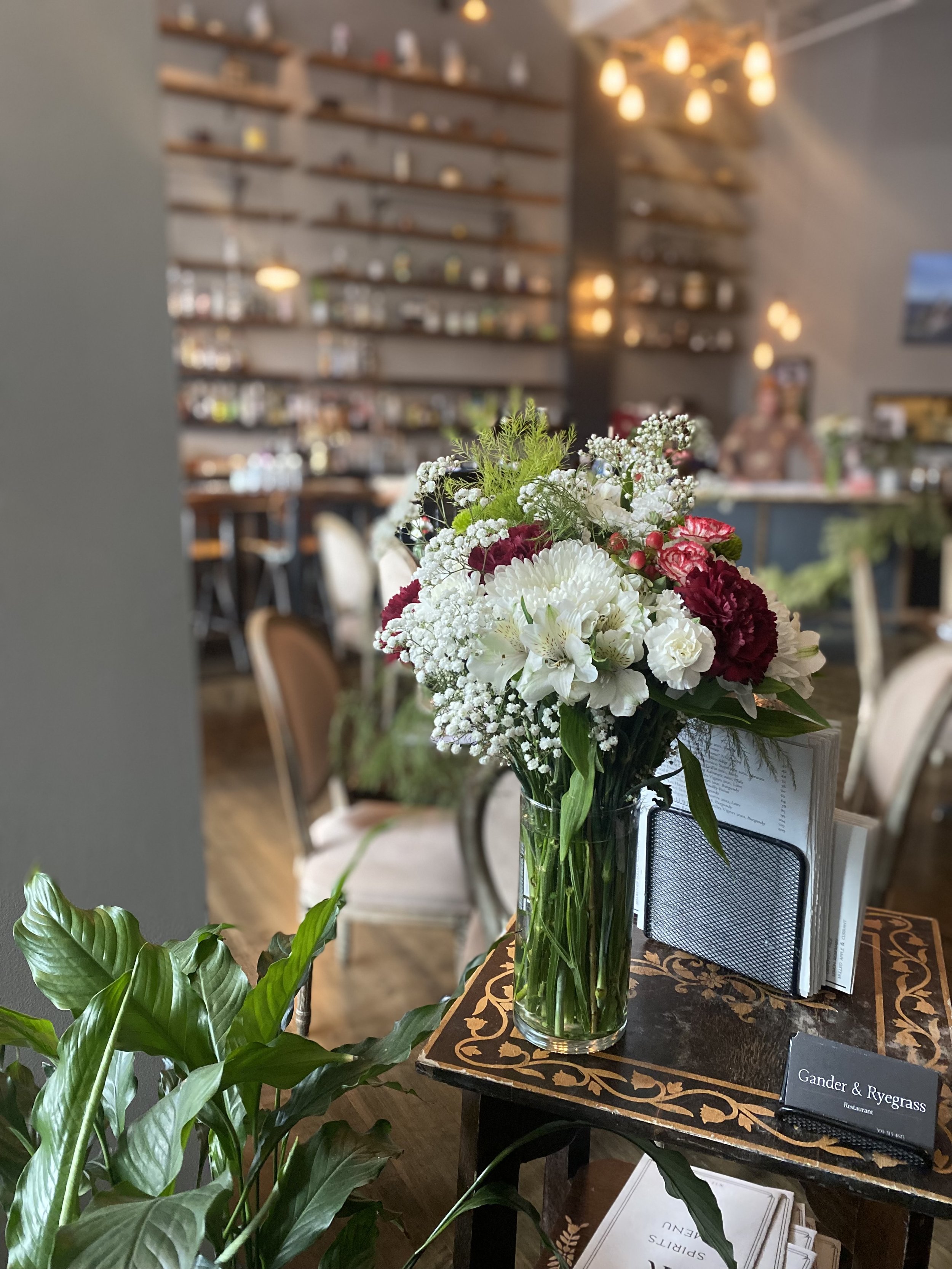 A vase of mixed white and red flowers on a table in a cozy restaurant with blurred background, shelves on the wall, and warm lighting.