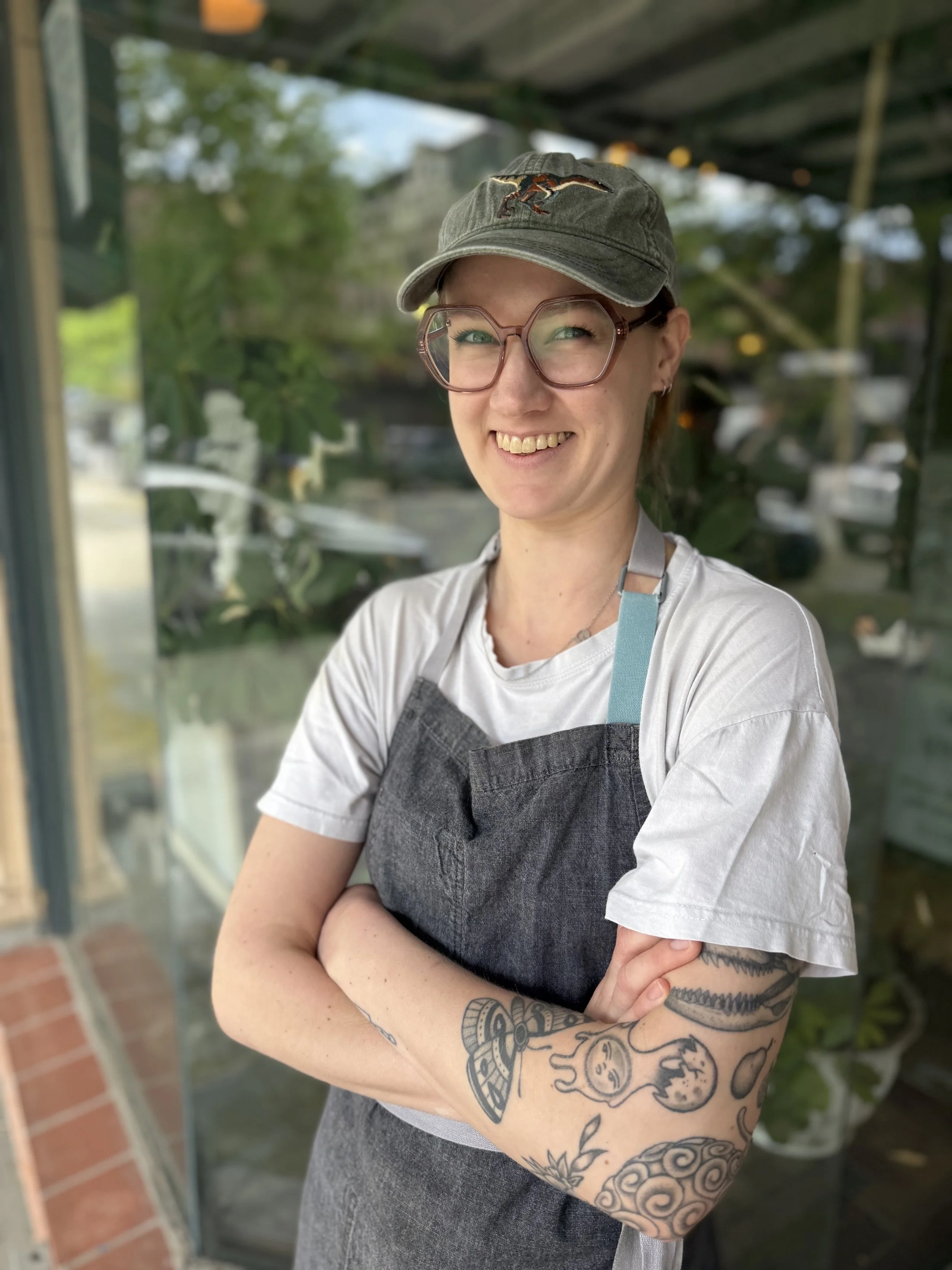 A smiling woman with tattoos on her arm, wearing glasses, a gray baseball cap, a white T-shirt, and a gray apron, stands outdoors with crossed arms.