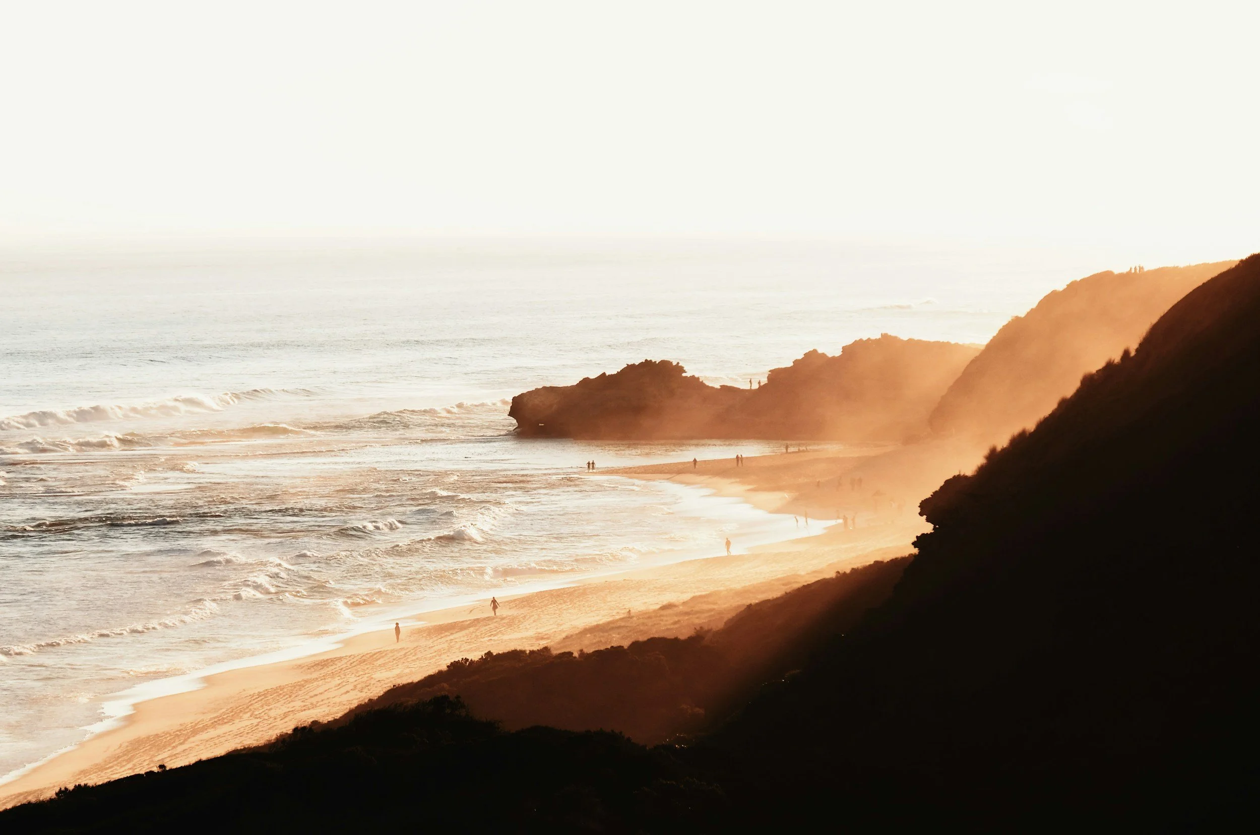 A scenic view of a beach at sunset with cliffs on the right, people walking along the shoreline, and waves crashing on the sand.