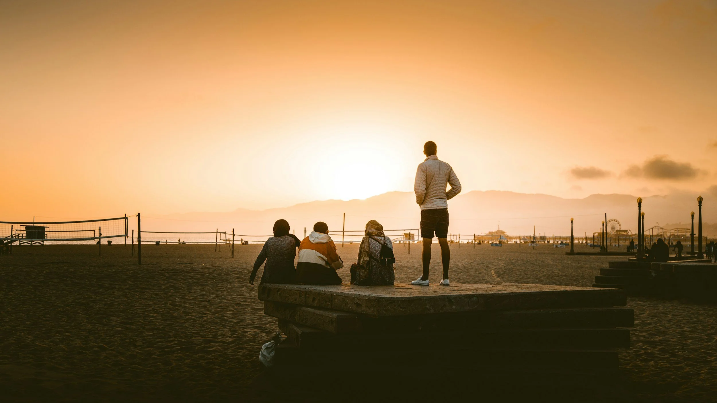People sitting and standing on a beach at sunset, with a ferris wheel in the background.