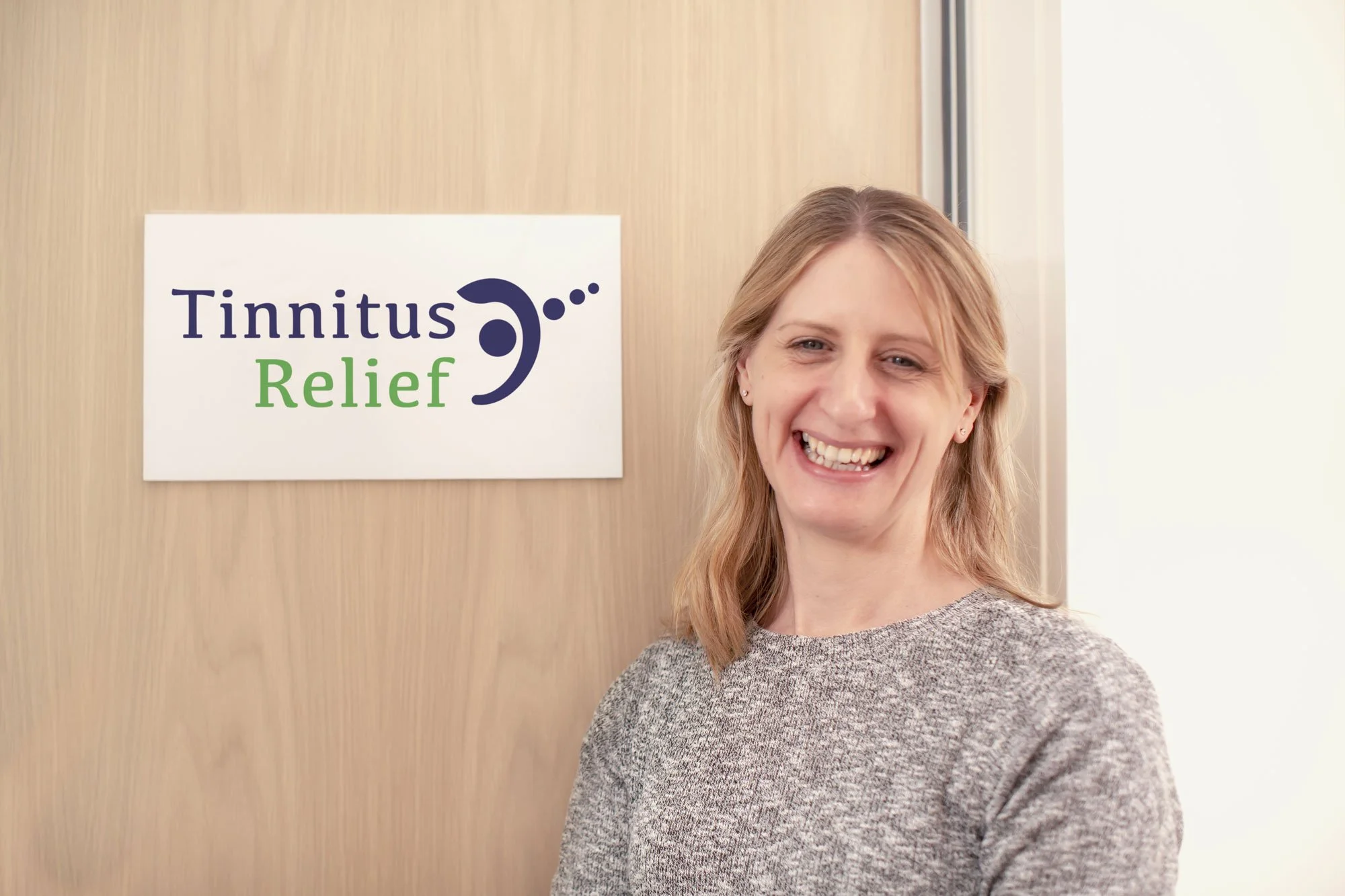 A woman smiling stands in front of a sign that reads 'Tinnitus Relief' with a stylized ear logo, in an office setting.