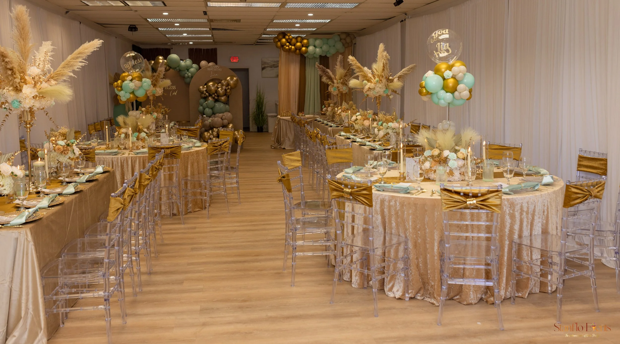 Decorated banquet hall with gold and pastel balloon arrangements, floral centerpieces, transparent chairs with gold bows, and long tables set for an event.