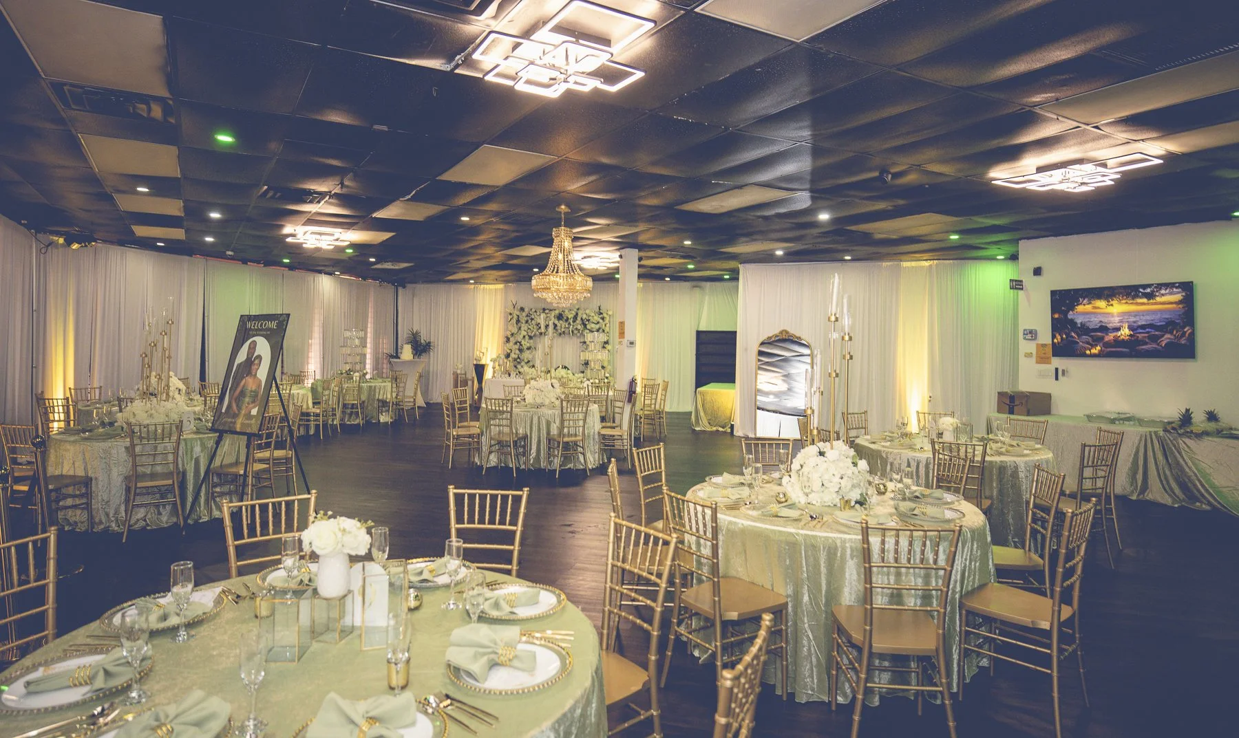Elegant banquet hall decorated for an event with round tables covered in green and gold tablecloths, adorned with white floral centerpieces, gold chairs, and a chandelier hanging from the ceiling. A