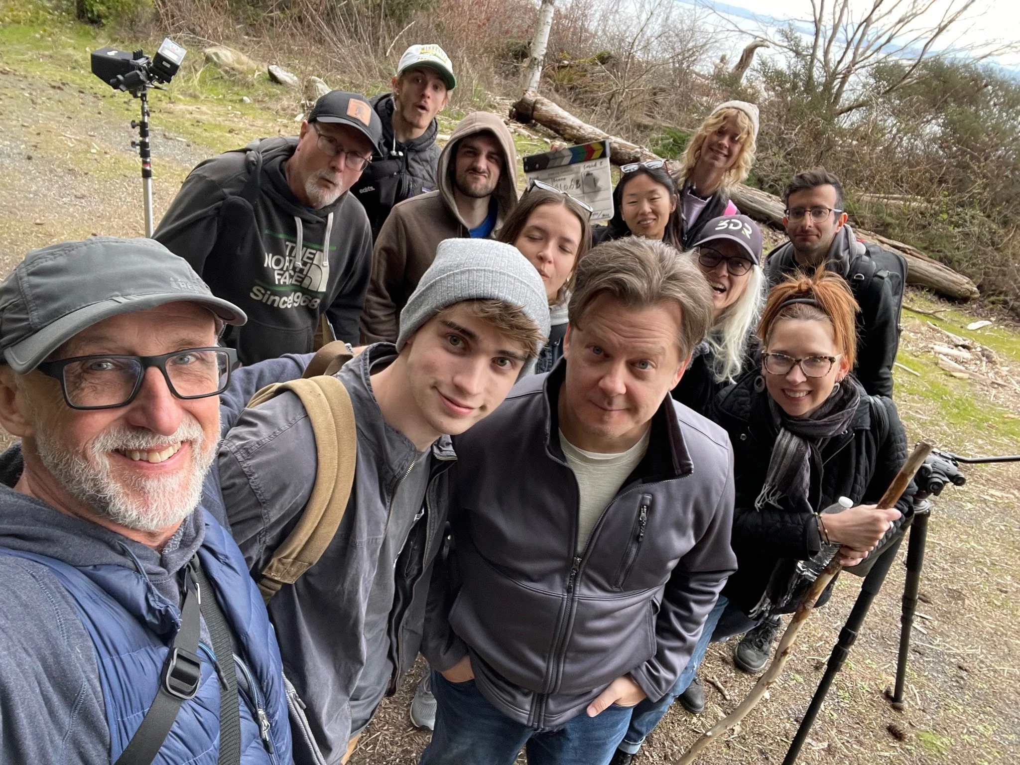 Group of people outdoors in a forest, taking a selfie. Some hold equipment like tripods and cameras, and the background includes trees and logs.