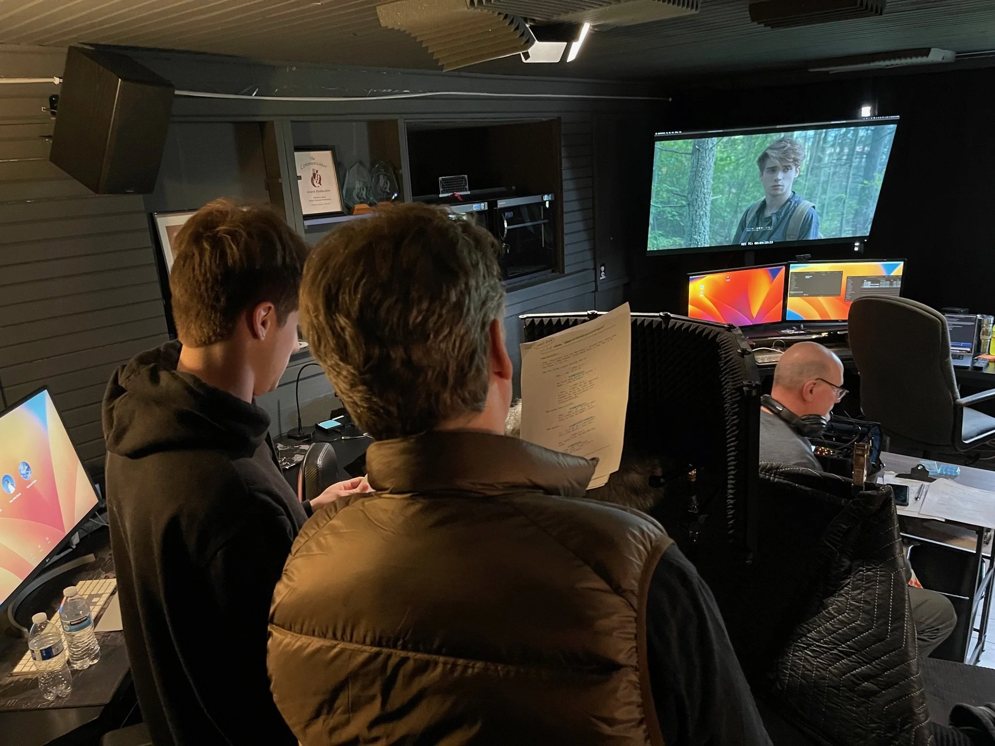Group of people in a room with multiple computer monitors, one person wearing headphones, a large screen displaying a scene from a movie or video, and another person holding a script or notes.