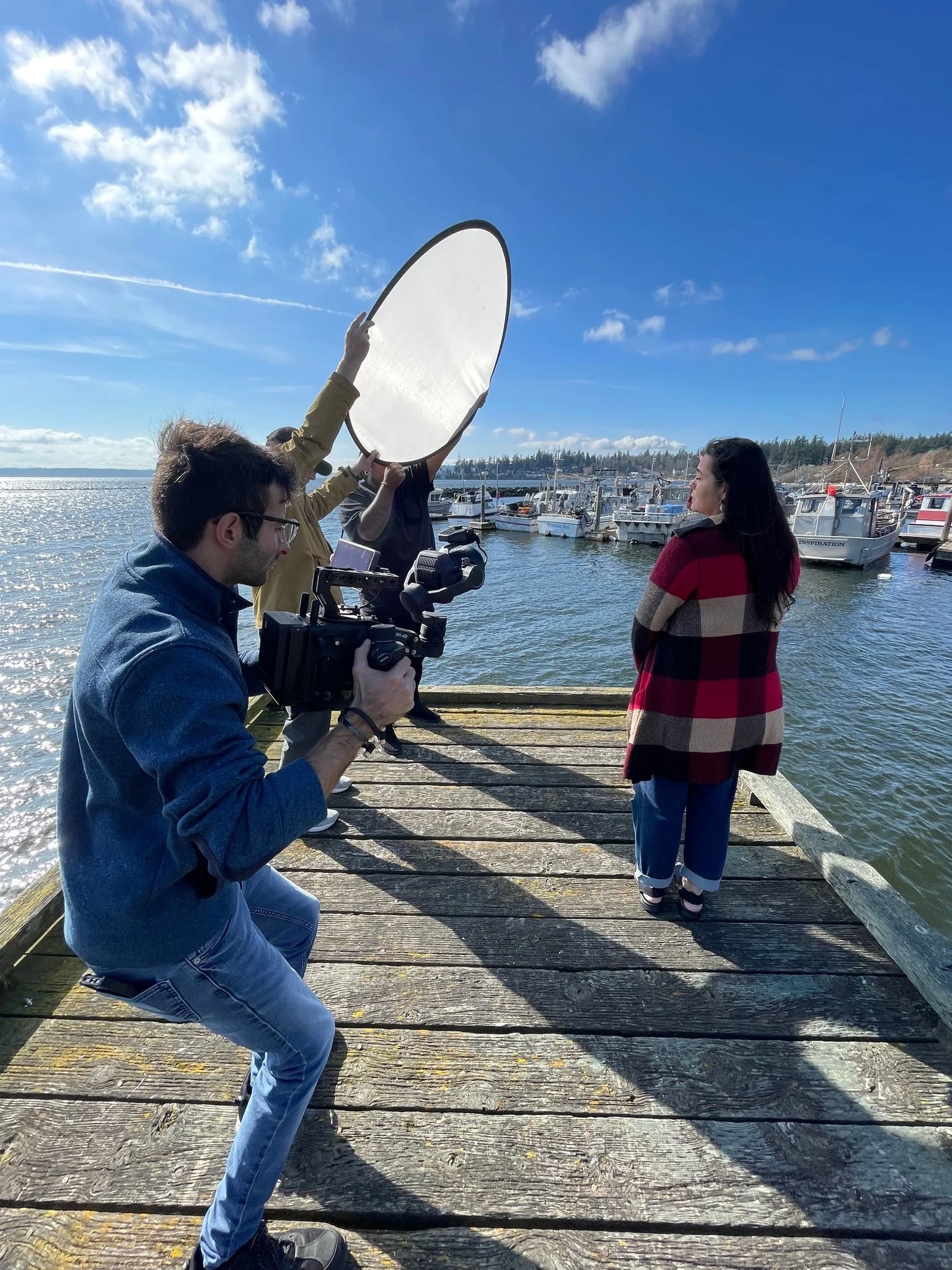 A woman standing on a wooden dock by the water, having her photo taken by a photographer with a camera, while another person holds a reflector to reflect sunlight onto her. Several boats are docked in the background against a clear blue sky.