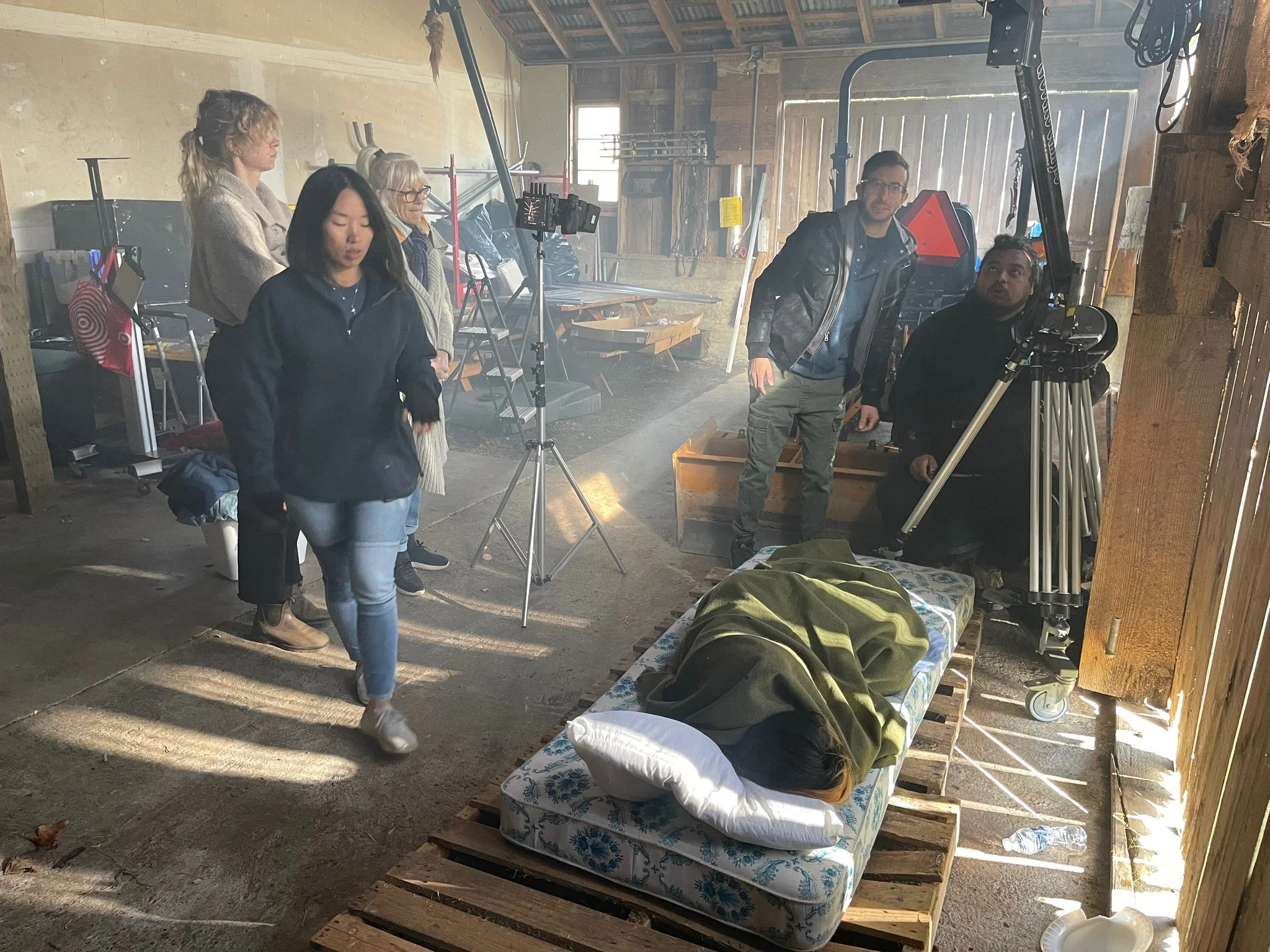 Film crew setting up a camera and lighting in a rustic barn, with a person lying on a pallet bed covered with a blanket, nearby.
