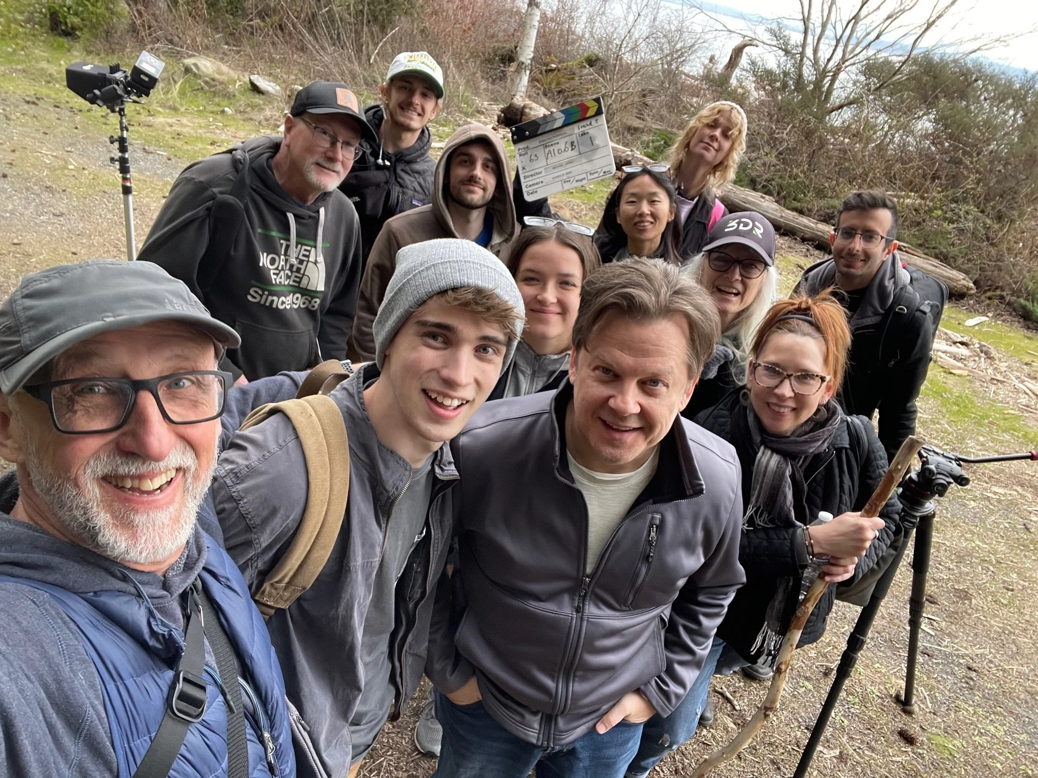 Group of people taking a selfie outdoors in a wooded area with a film crew clapperboard in the background.