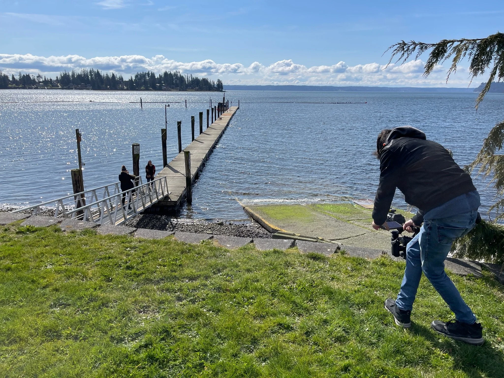A man operating a camera on a grassy area near a dock leading into a body of water with a few people standing at the end of the dock, and an island with trees in the background under a partly cloudy sky.