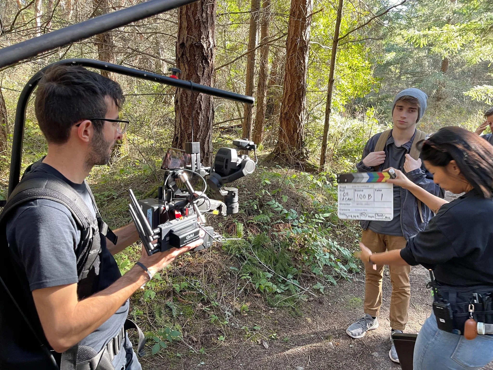 Filmmaking crew filming in a forest, with one person holding a camera and another holding a clapperboard.