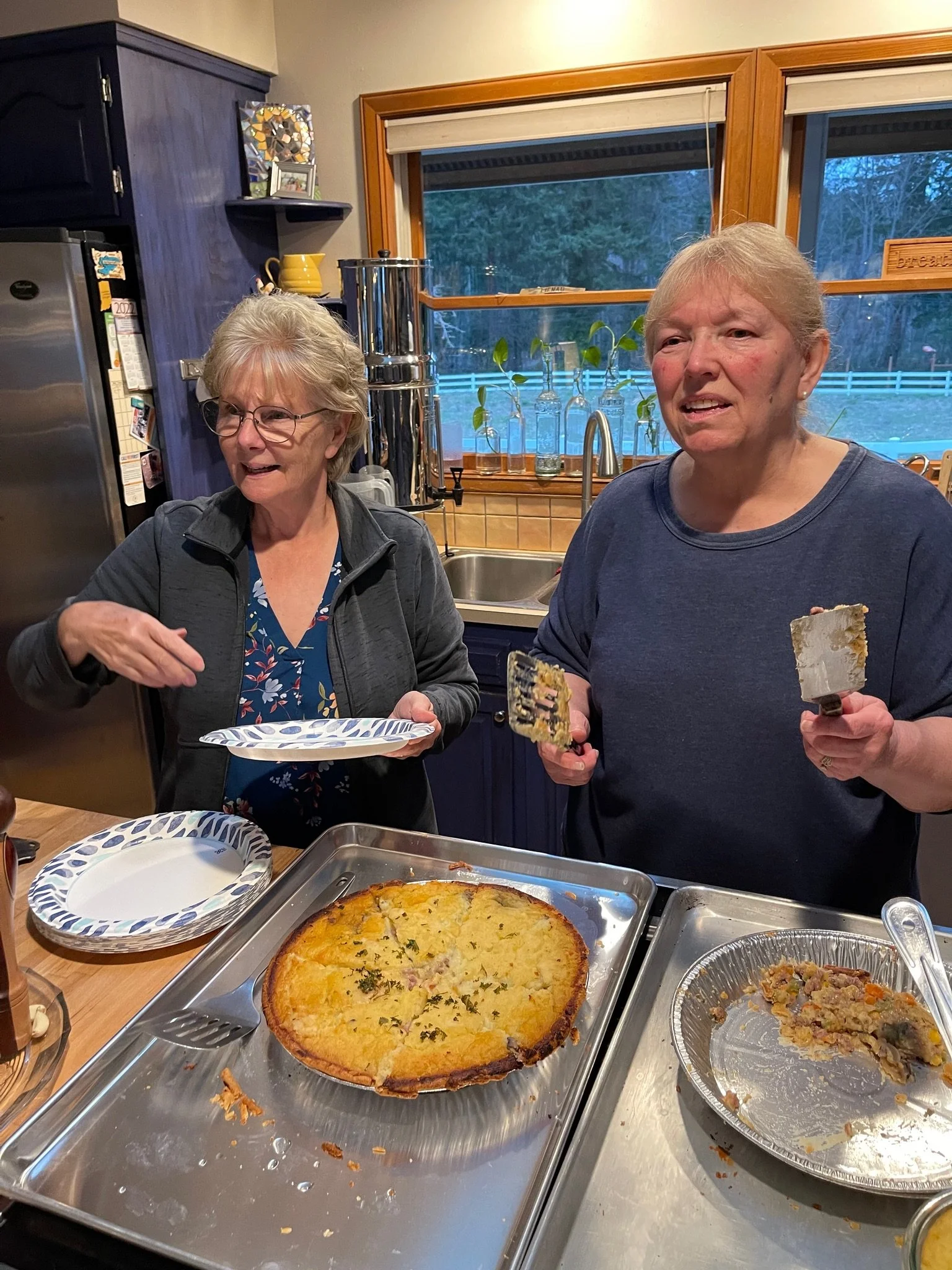 Two women standing at a kitchen counter with pizza and food, chatting. One woman is holding a pizza cutter, the other is holding a slice of pizza on a spatula. Behind them are kitchen cabinets, a window, and a home interior.