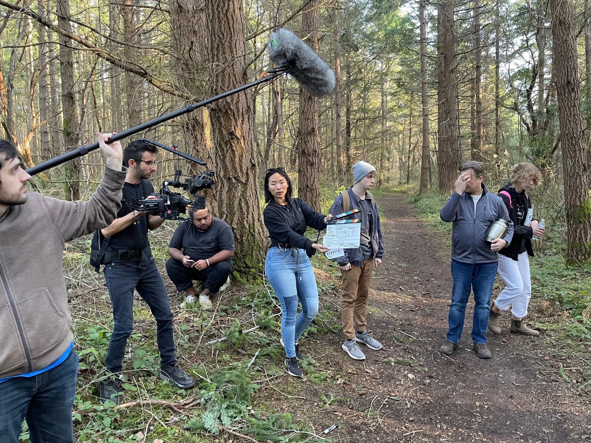 Filming crew setting up a scene in a wooded area with trees and a dirt trail, including a person holding a boom microphone.
