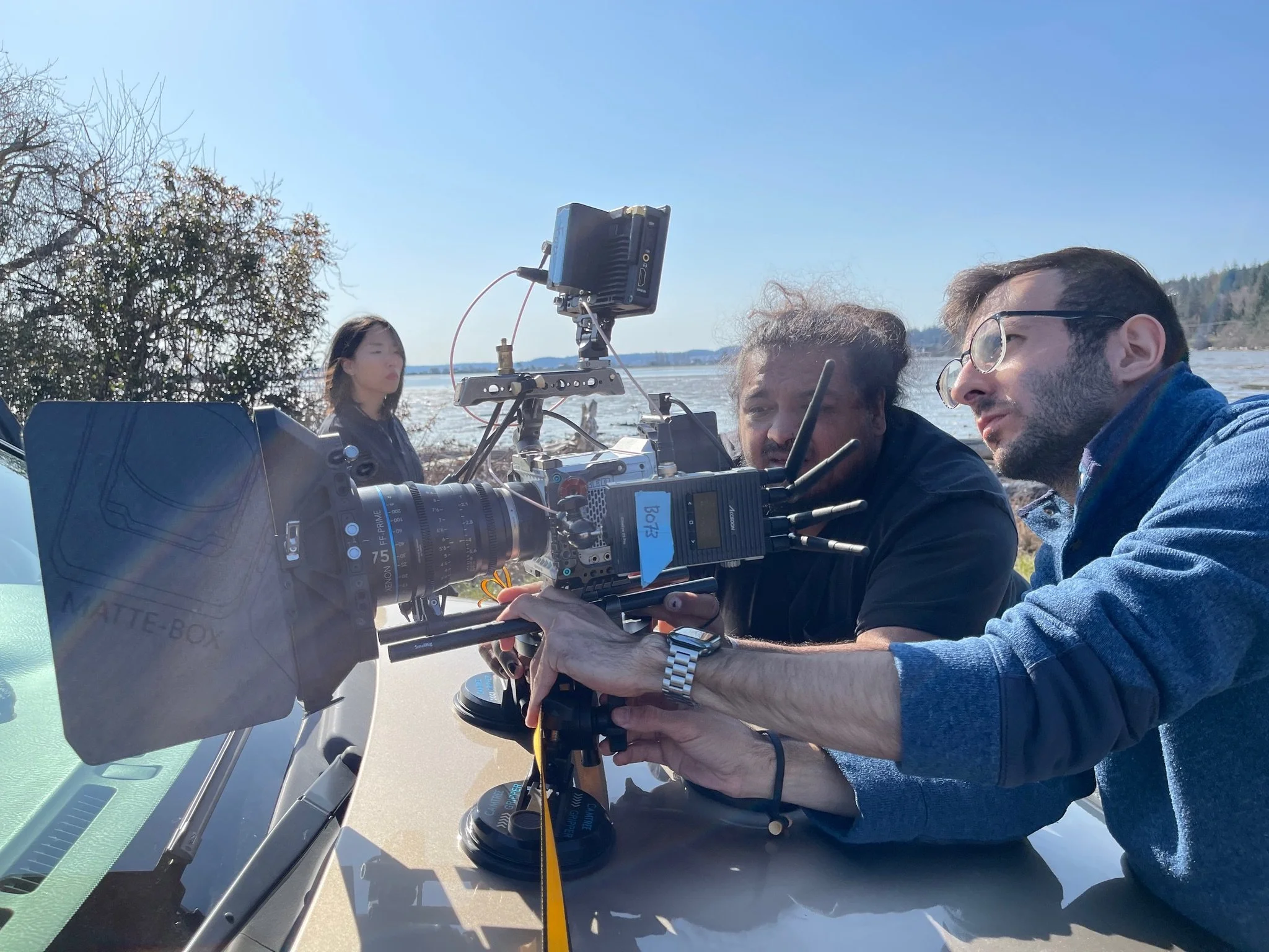 Two people operating a camera with a third person in the background near a body of water on a sunny day.