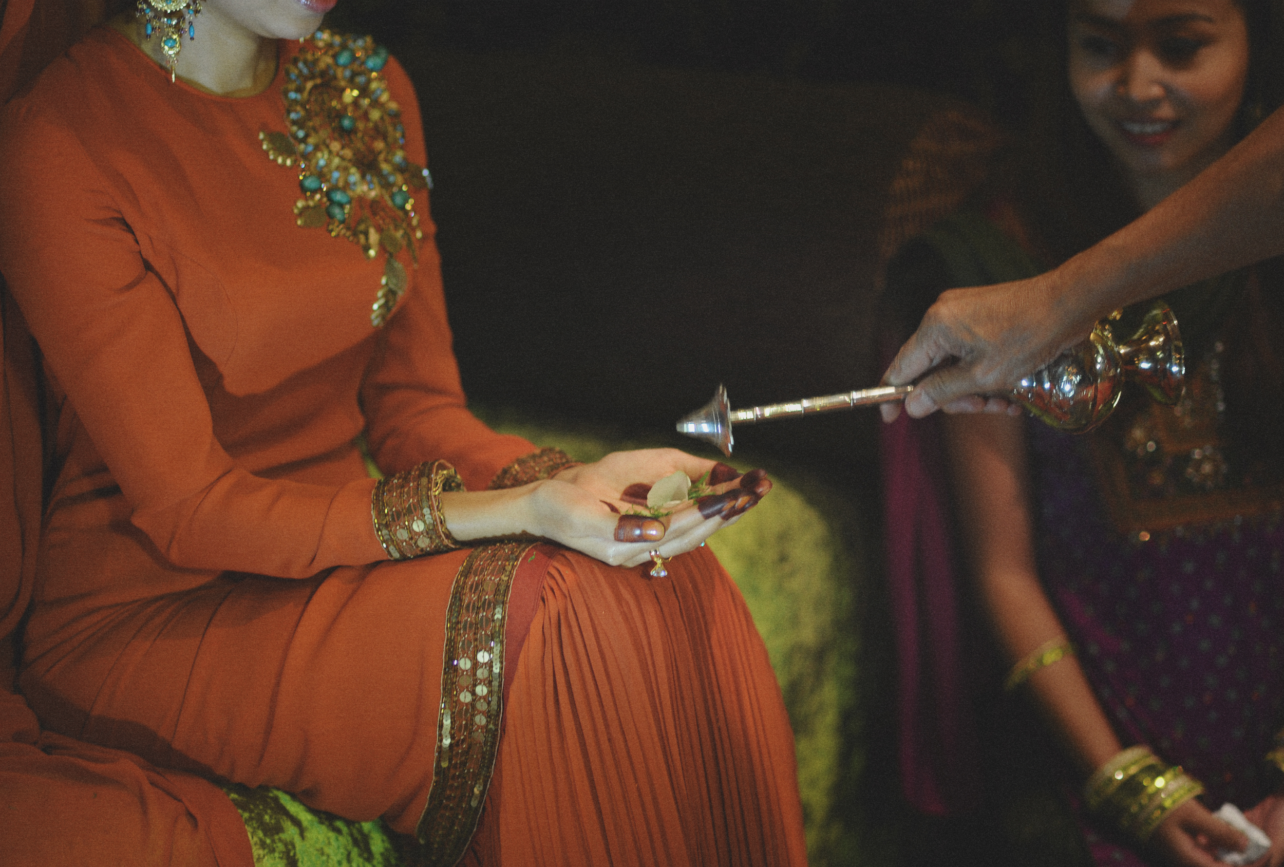 A woman dressed in traditional Indian attire receiving a religious blessing with a brass lamp during a ceremony. Another woman looks on smiling.