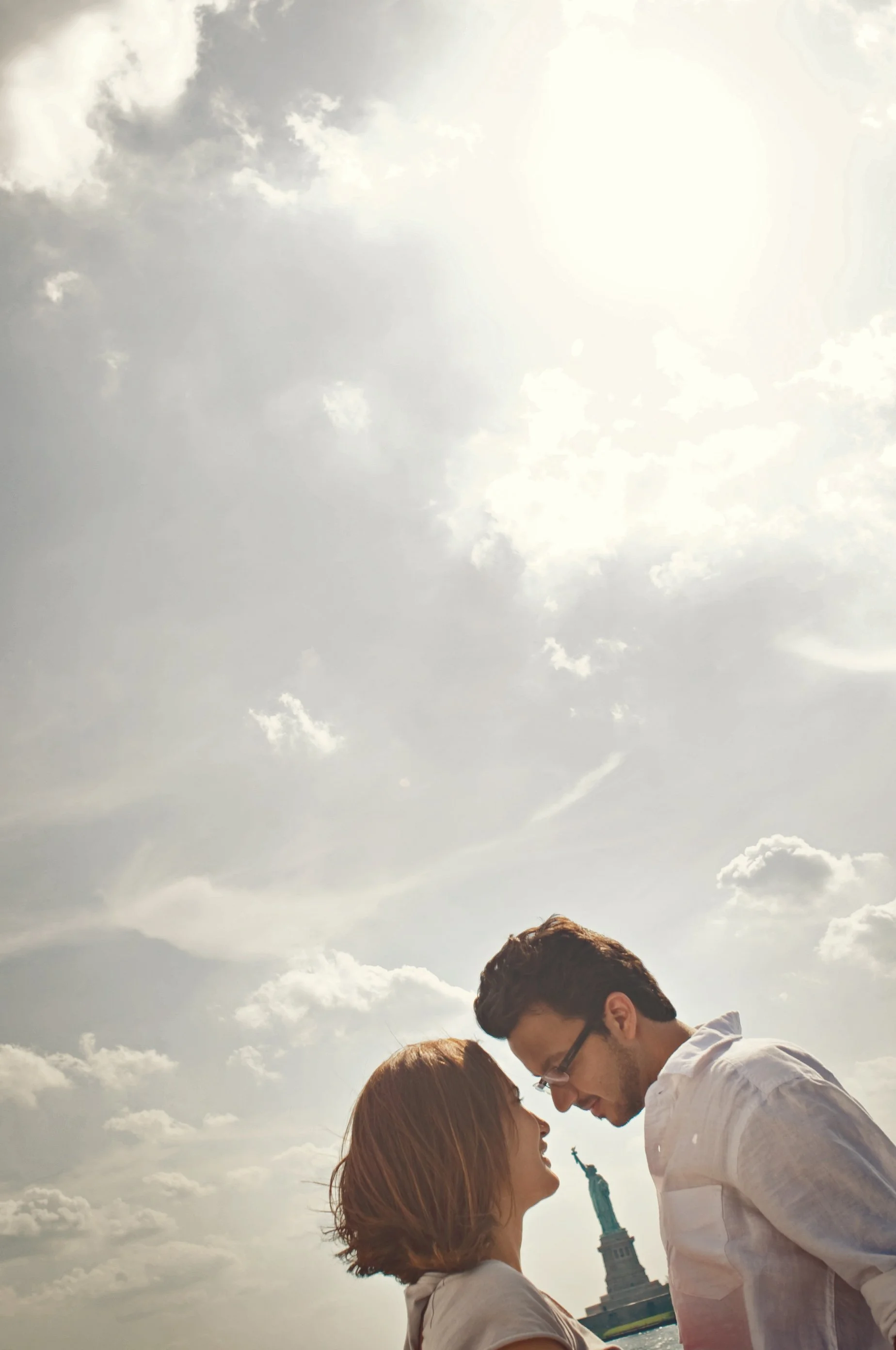 A couple standing outdoors with the Statue of Liberty in the background, looking at each other, under a cloudy sky.