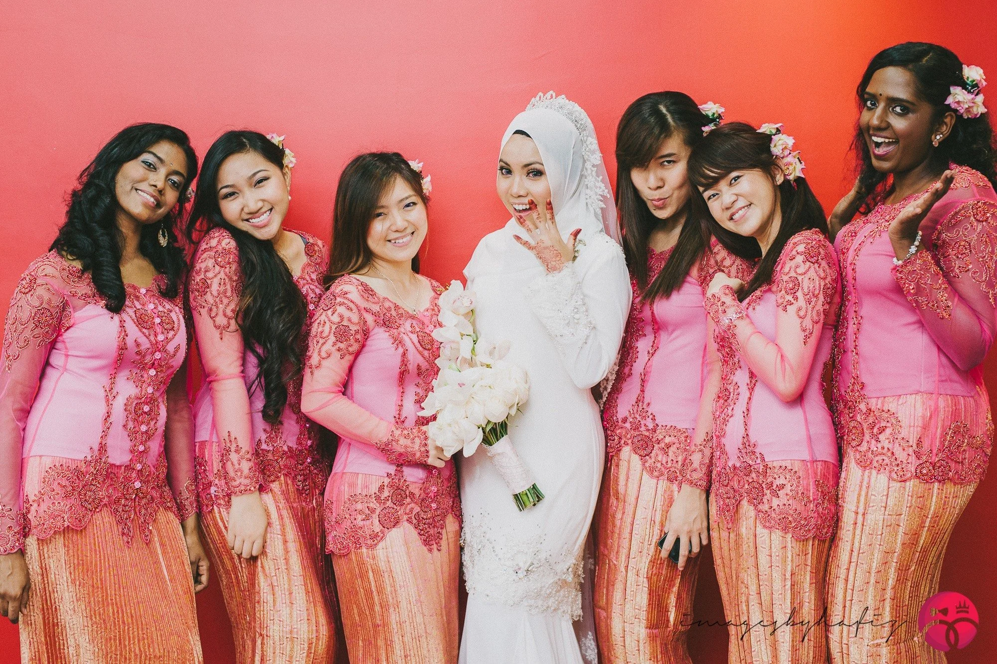 A bride in a white wedding dress holding a bouquet stands with seven women in pink traditional dresses against a pink background.
