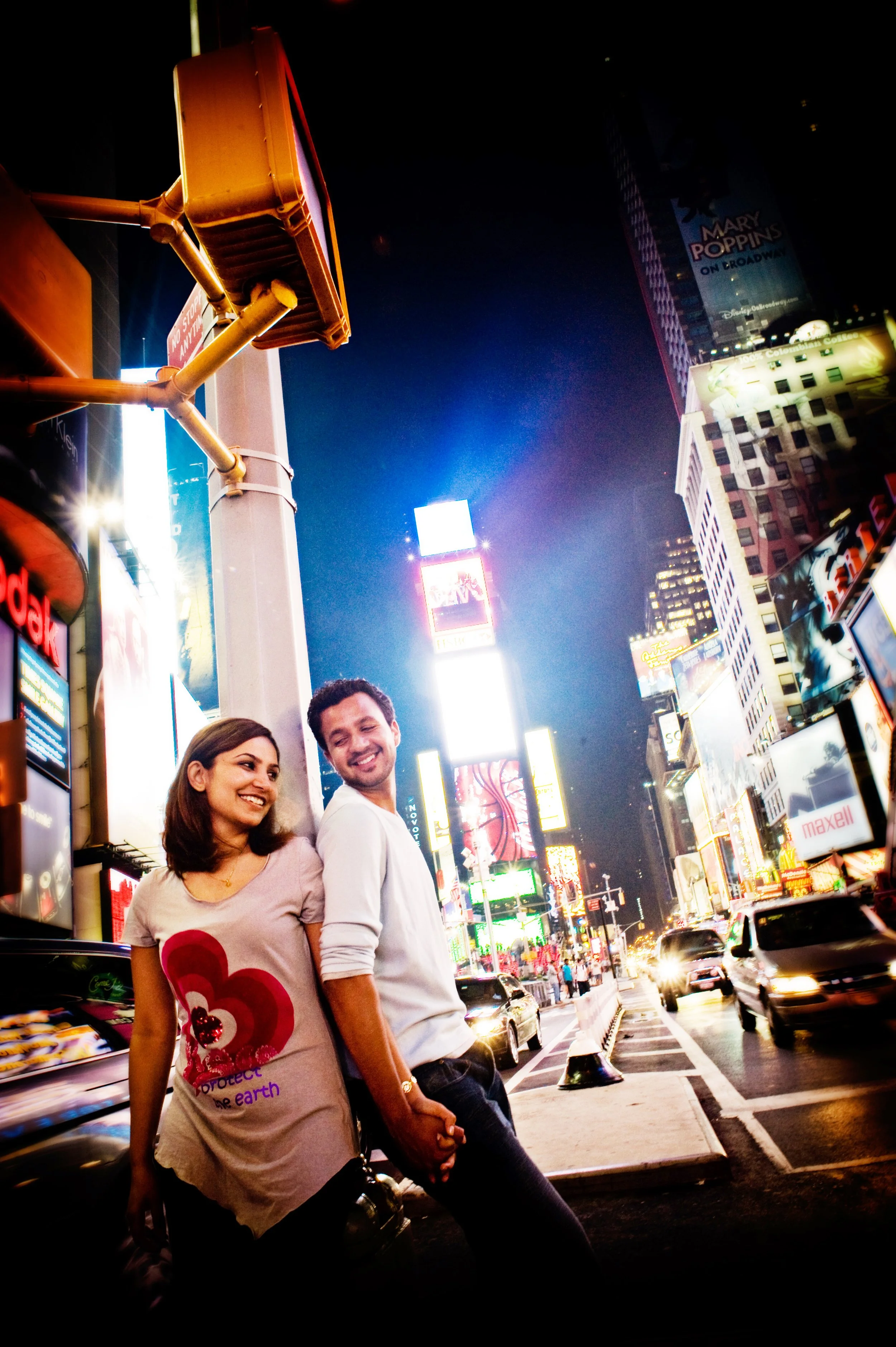 A smiling couple holding hands and walking in Times Square at night, surrounded by bright billboards and illuminated signs.