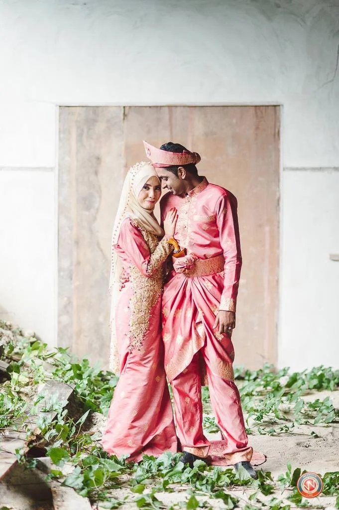 A couple dressed in traditional pink and gold attire, posing together during a celebration or wedding, with greenery and rustic background elements.