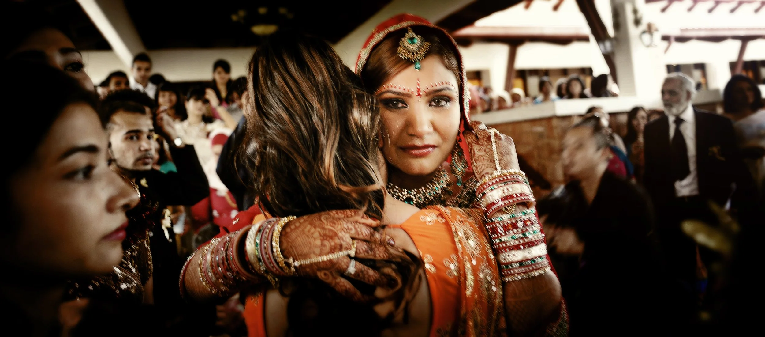 Two women hugging at a celebration, one dressed in traditional Indian attire with jewelry, and the other woman with henna on her hands surrounded by people in a festive setting.