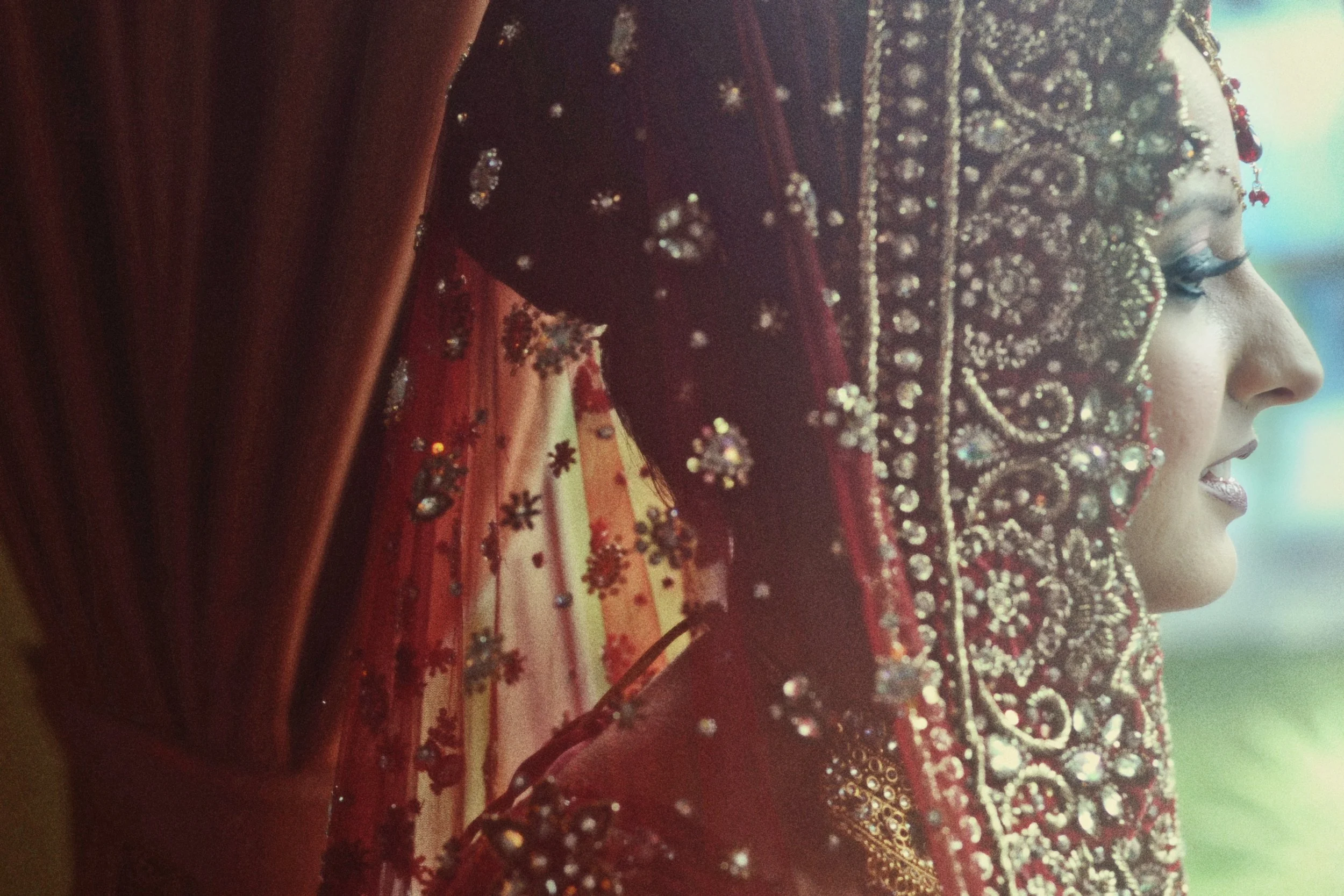 A woman dressed in traditional South Asian bridal attire with an elaborately embroidered and jeweled red dupatta covering her head, showcasing detailed beadwork and sequins, with her profile visible and a soft focus background.