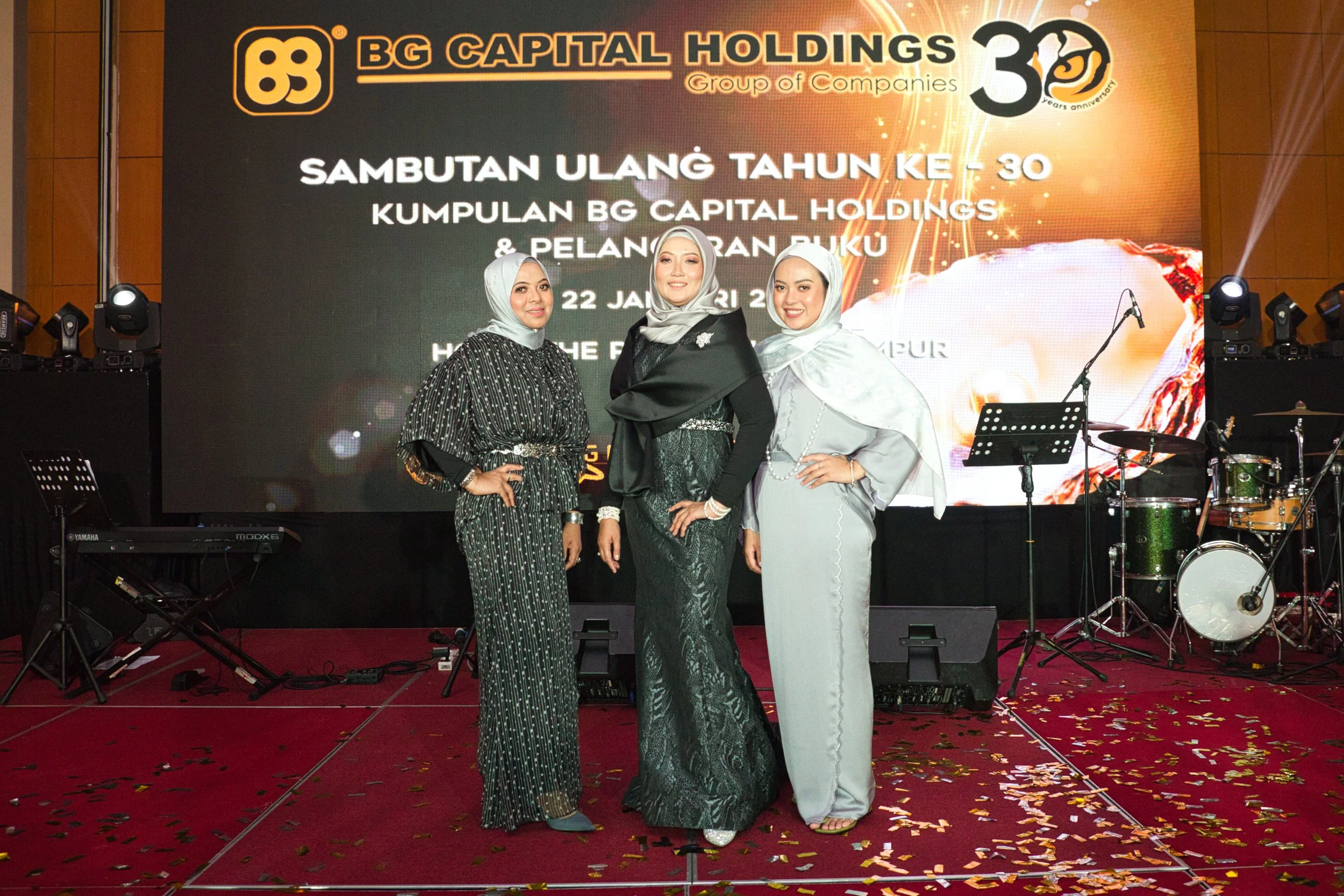 Three women wearing elegant traditional attire and headscarves, standing on a stage with a large screen behind them displaying text celebrating the 30th anniversary of BG Capital Holdings, with musical instruments and stage lights present.