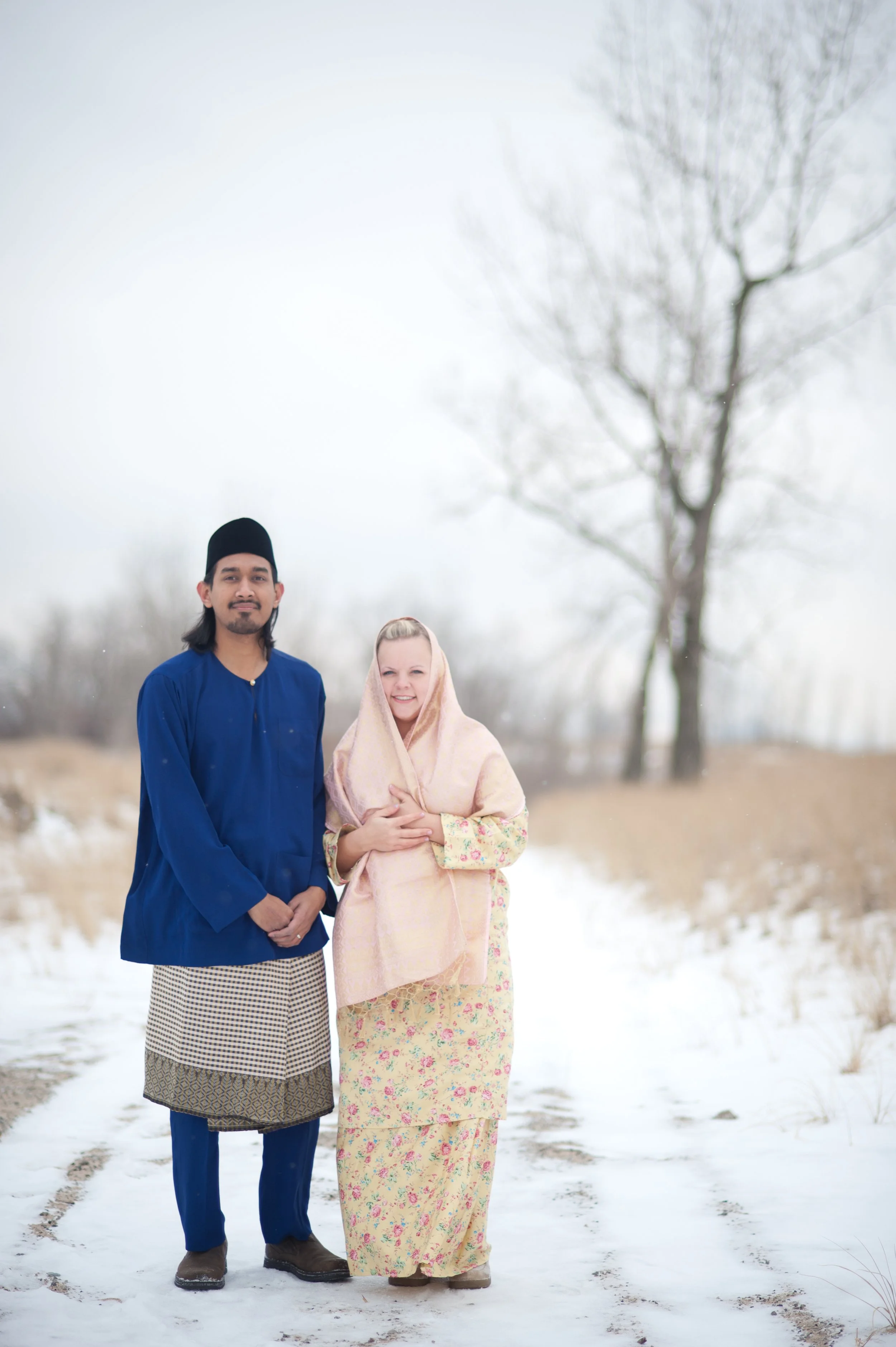 A man and a woman standing together on a snowy path outdoors, dressed in traditional clothing, with leafless trees in the background.