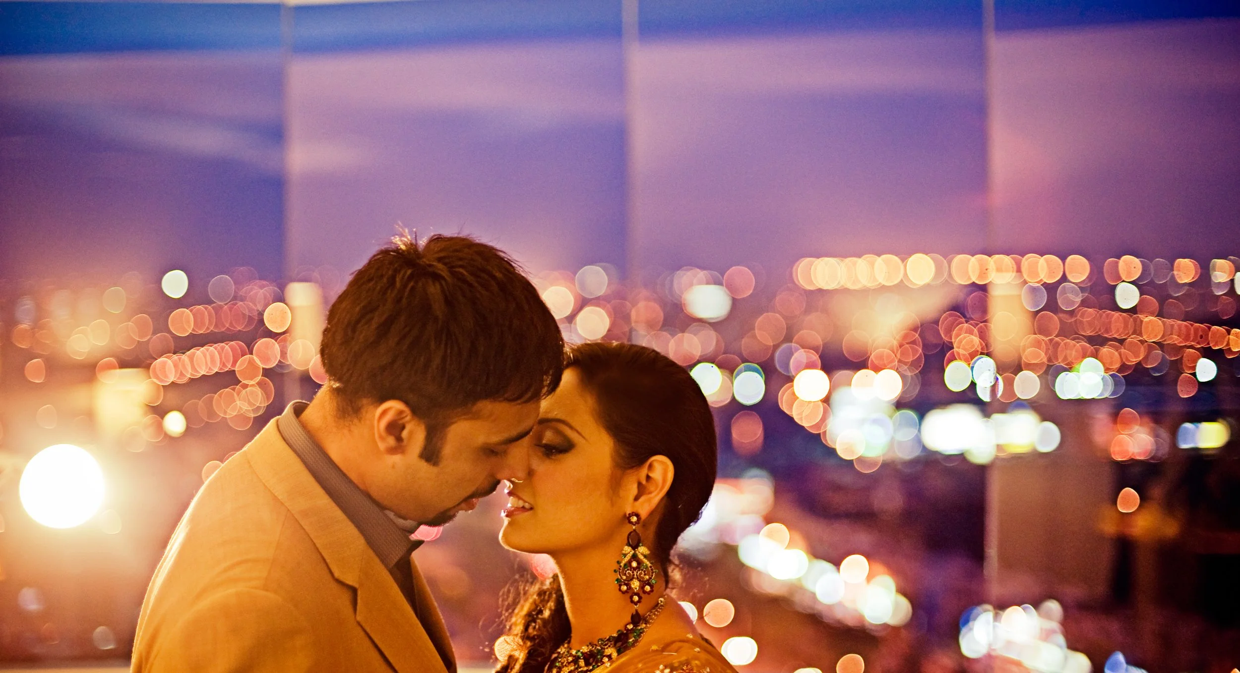 A romantic moment between a man and a woman with their foreheads touching, at night on a rooftop with blurred city lights in the background.