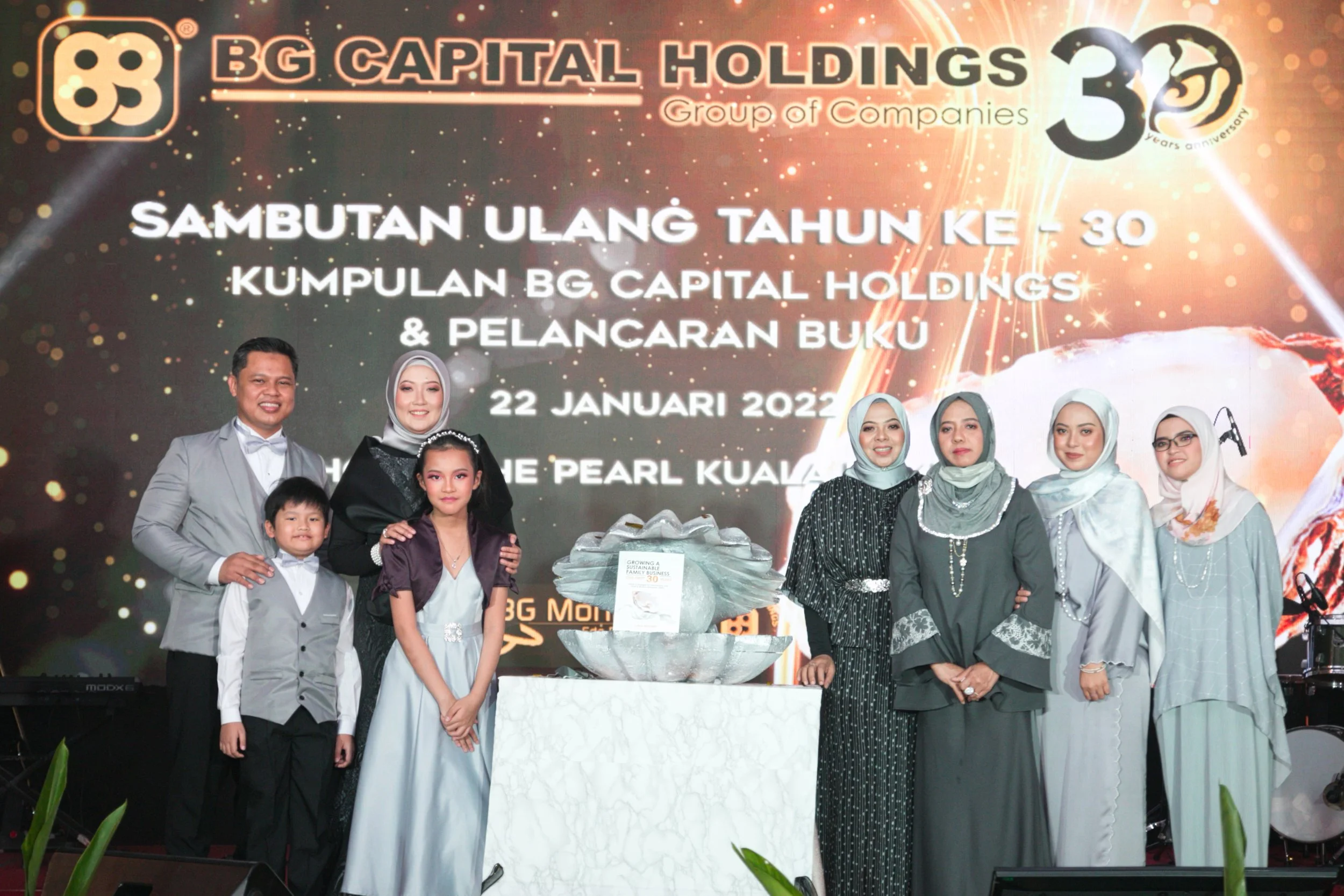 Group of people celebrating the 30th anniversary of BG Capital Holdings with a backdrop displaying event details in Malay, including date and location. Four adult women, a man, and two children are standing on stage, some dressed in formal attire and