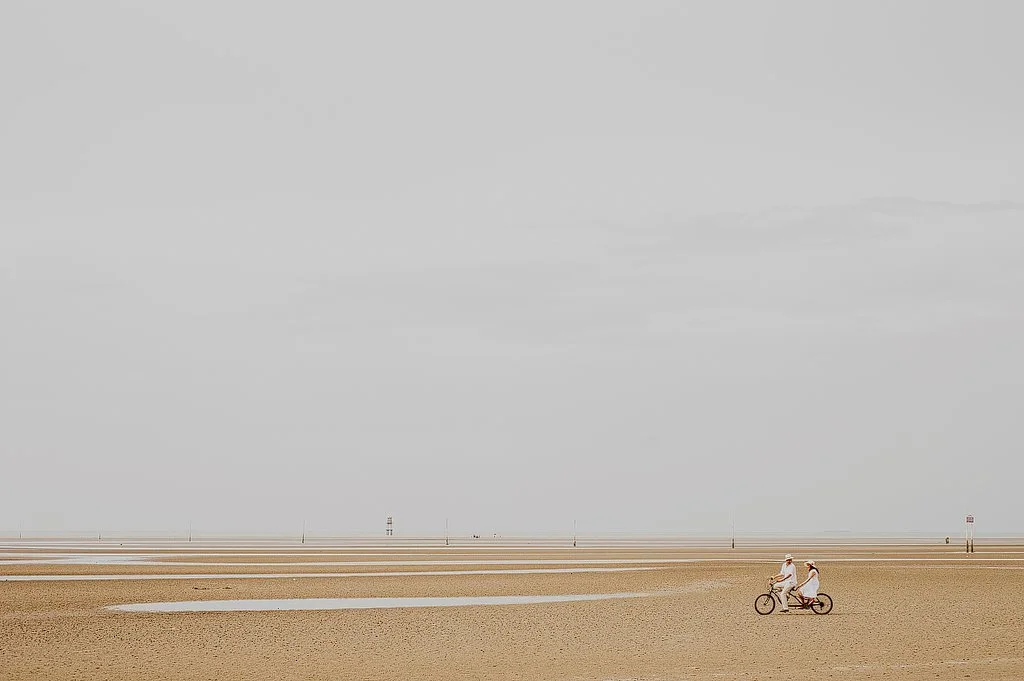 Two people riding a tandem bicycle on a sandy beach against a cloudy sky.