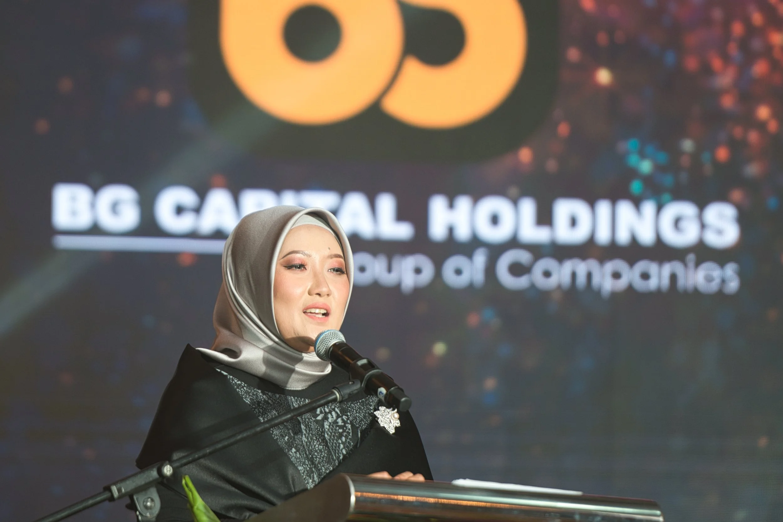 A woman wearing a gray hijab and black outfit speaking at a podium during an event. Behind her is a large screen displaying the logo and name of BG Capital Holdings, a group of companies.
