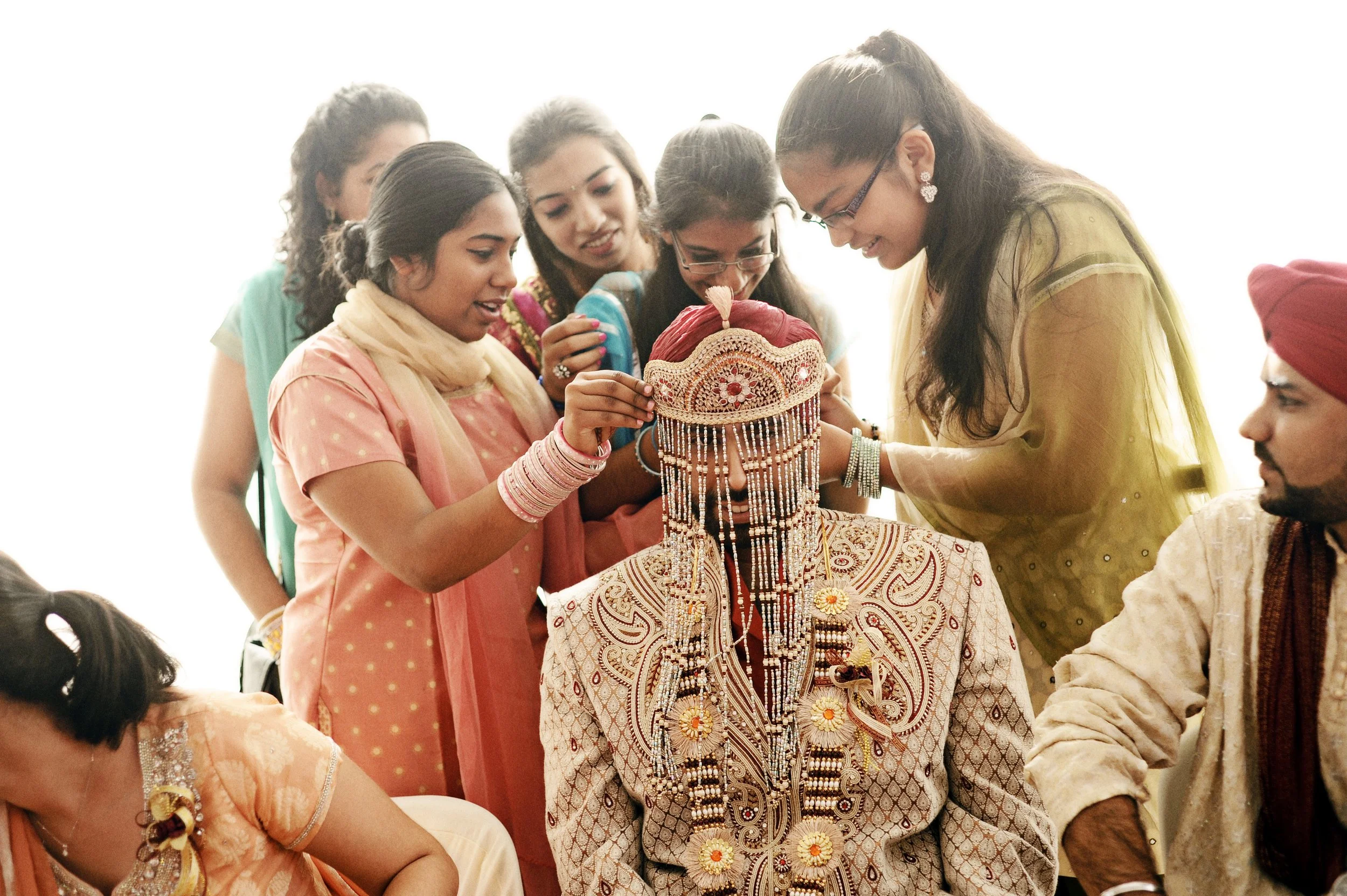 Group of women dressed in traditional Indian attire participating in a wedding ceremony, placing jewelry on a groom's head.