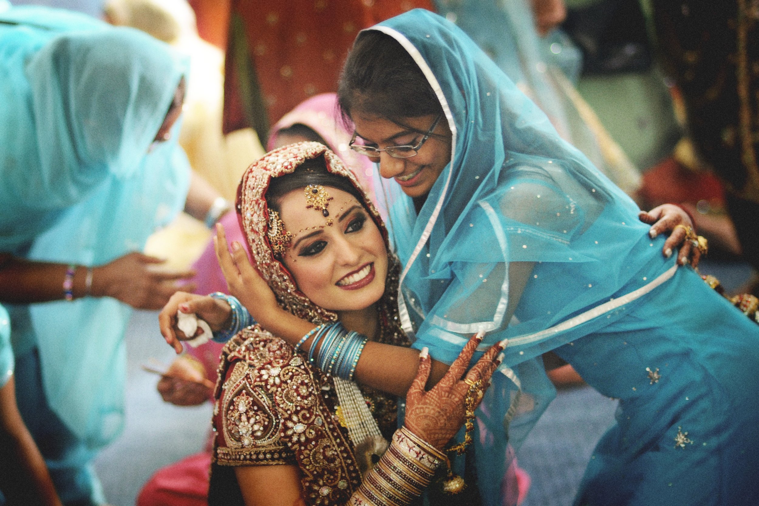 A bride in traditional Indian wedding attire, decorated with jewelry and henna, smiling as a woman in a blue sari hugs her during a celebration.