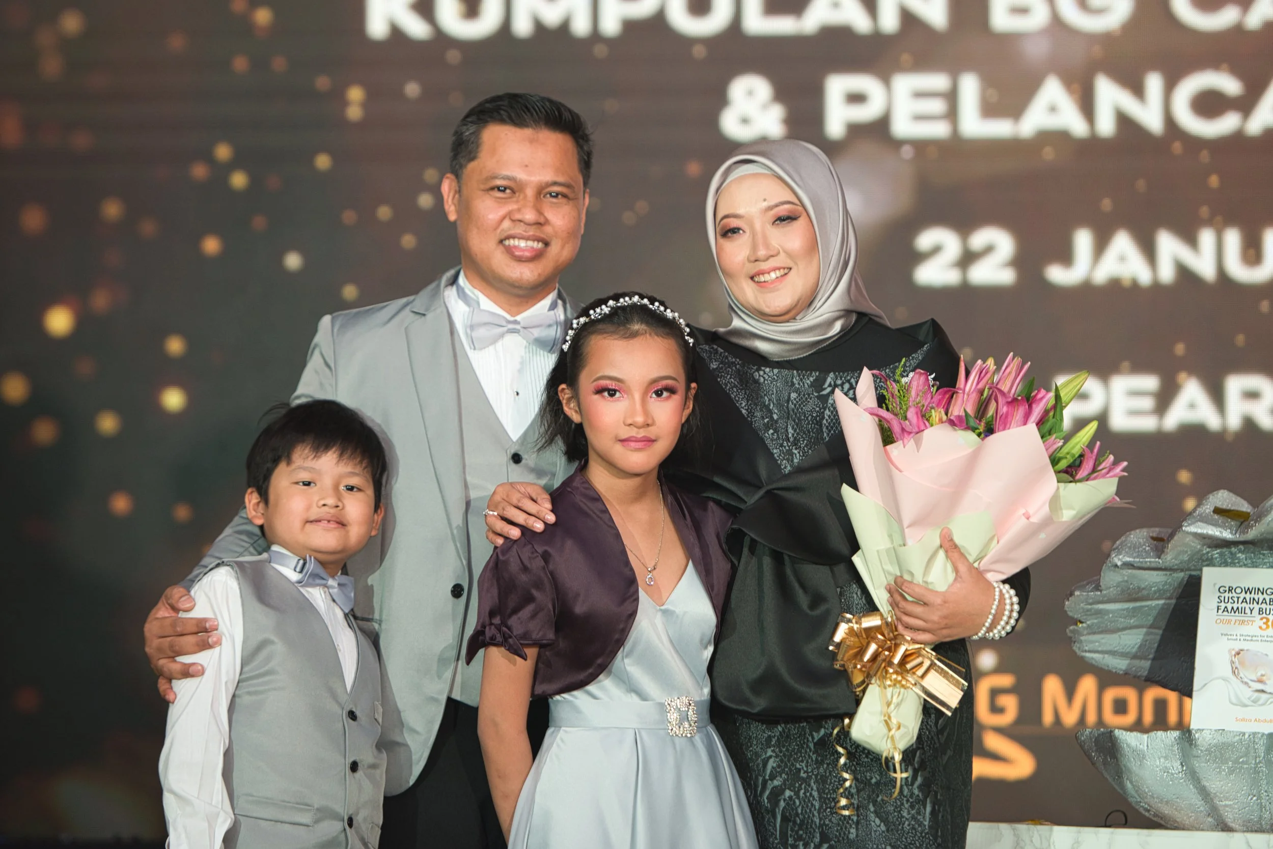 A family of five, including a man, woman, two girls, and a boy, gathered on a stage at an event, with the woman holding a bouquet of pink lilies. The backdrop displays text and event details.