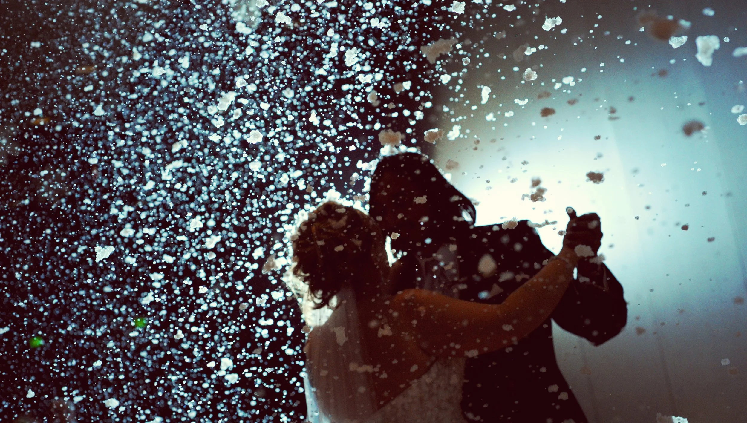 Silhouettes of a couple dancing, seen through water droplets or snowflakes on a glass surface, with a bright light source behind them creating a backlit effect.