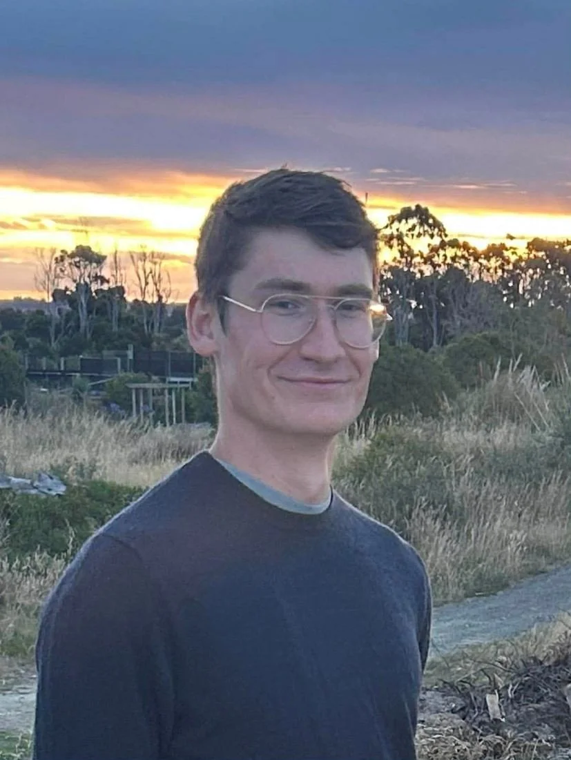 A young man with light brown hair sitting in a chair, wearing a black Adidas t-shirt, with a tattoo visible on his right arm, and a silver bracelet on his left wrist.