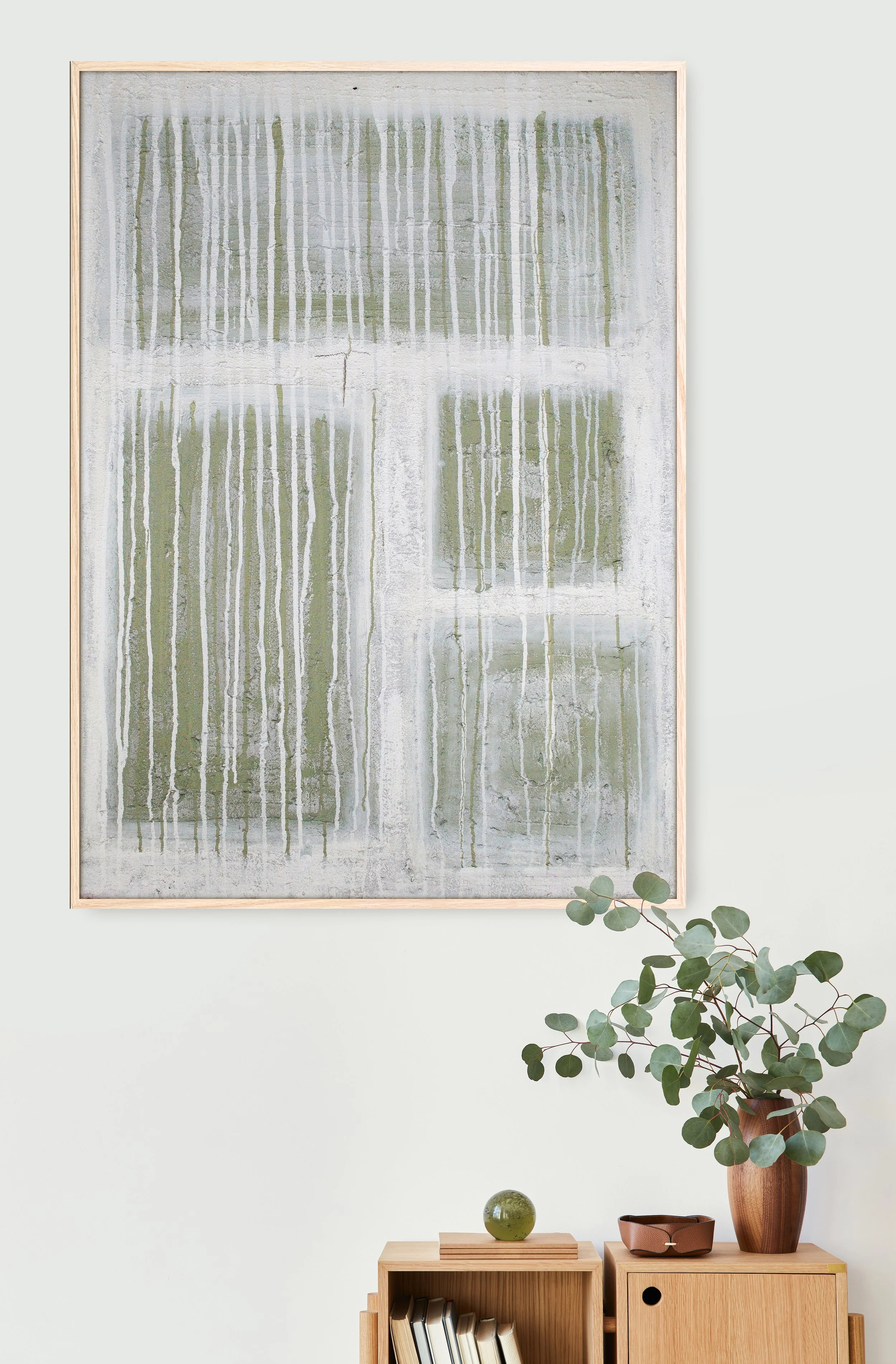 A minimalist interior decor with a large abstract painting featuring vertical streaks in shades of green, white, and gray. A wooden shelf with books, a green decorative object, a brown bowl, and a potted eucalyptus plant is positioned below the artwo