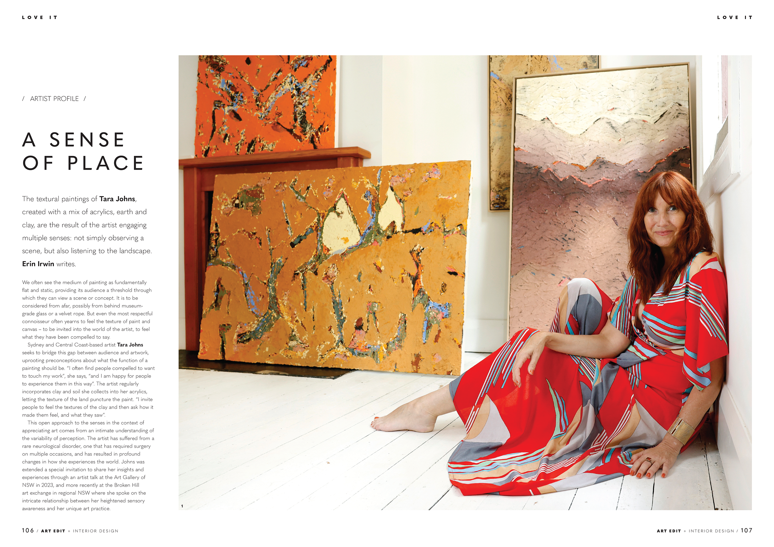 A woman with red hair and a colorful patterned dress sitting on the floor against a white wall in an art gallery. She is smiling and surrounded by abstract landscape paintings with textured brushstrokes and vibrant colors.