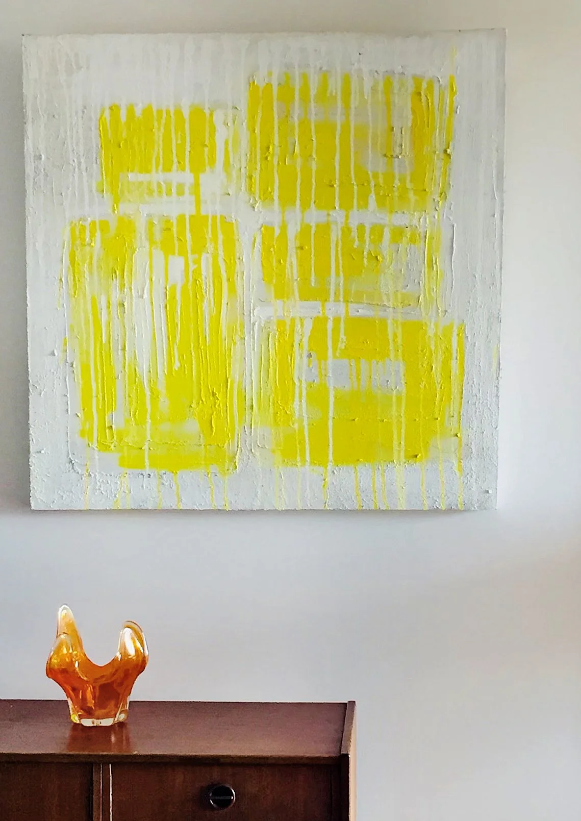 Abstract painting with yellow and white textures hanging on a wall above a wooden cabinet with an amber-colored decorative glass sculpture on top.