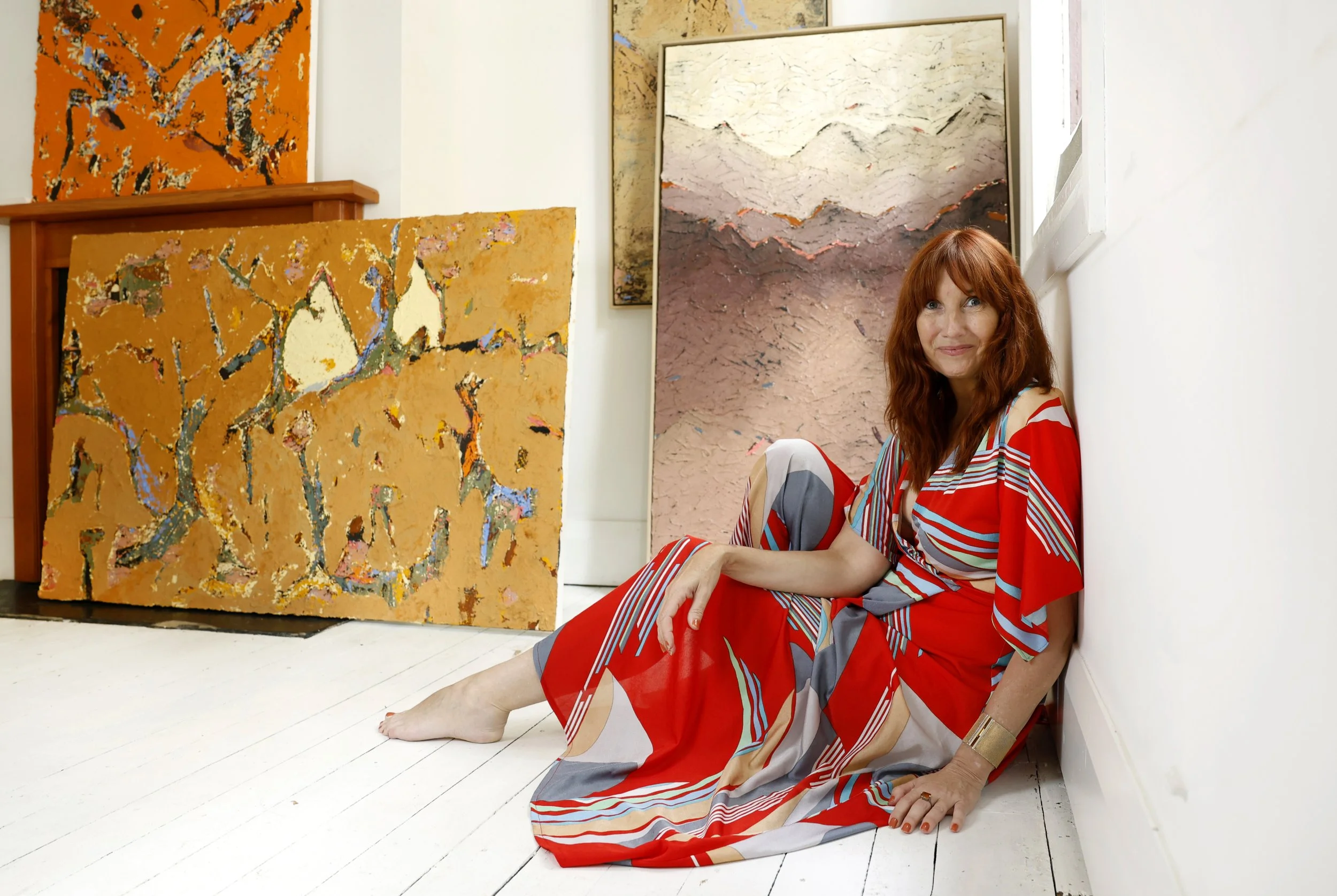 A woman with red hair in a colorful dress sitting on the white wooden floor of an art gallery, surrounded by abstract landscape-style paintings.